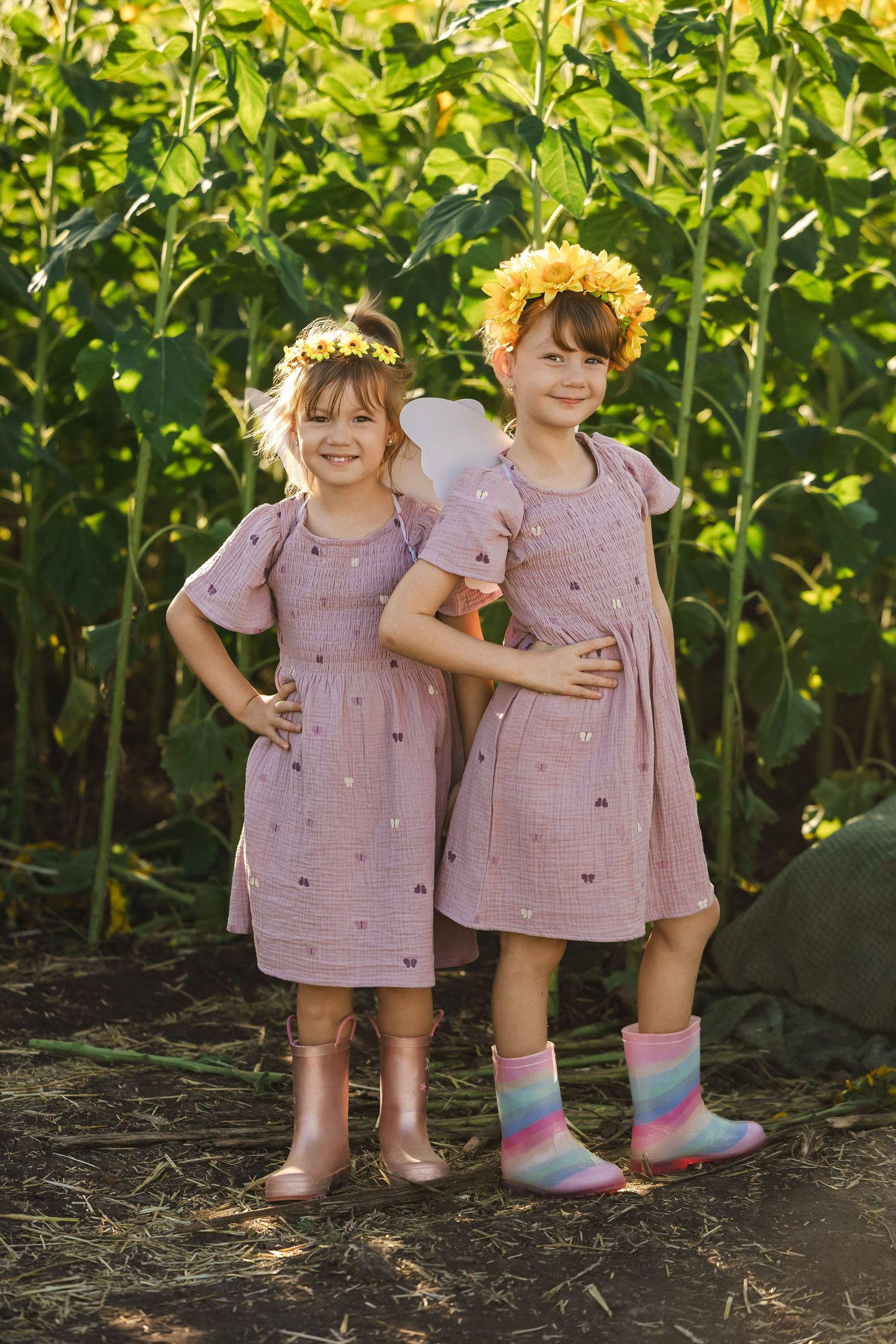 Two young girls in matching pink dresses and flower crowns pose together in a lush sunflower field.