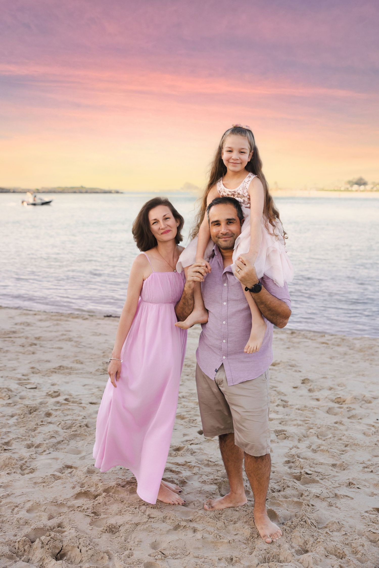 Family of three posing on sandy beach at sunset, mother in pink dress, father holding daughter on shoulders.