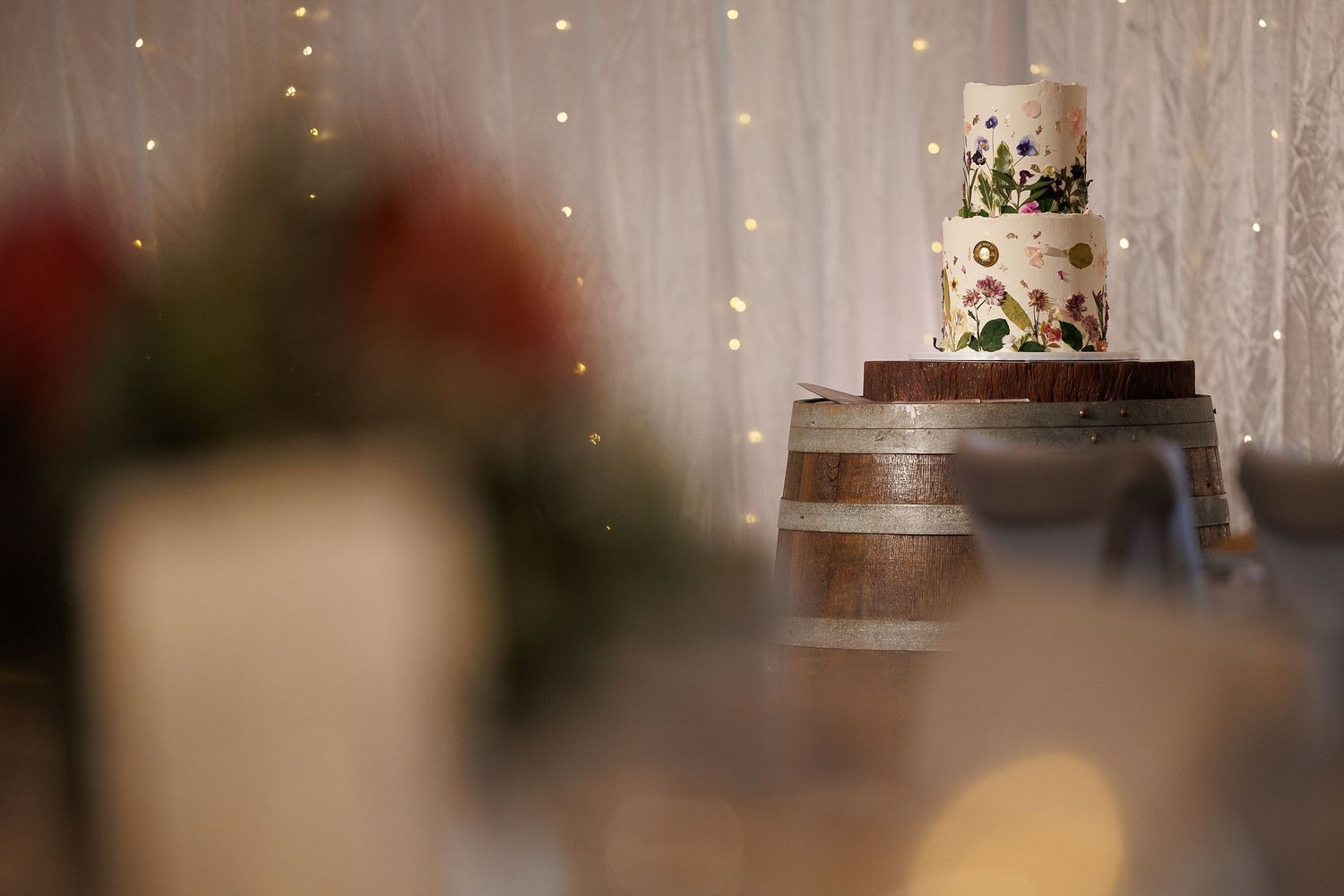 Elegant two-tier floral wedding cake displayed on a wooden barrel stand with twinkling fairy lights backdrop.