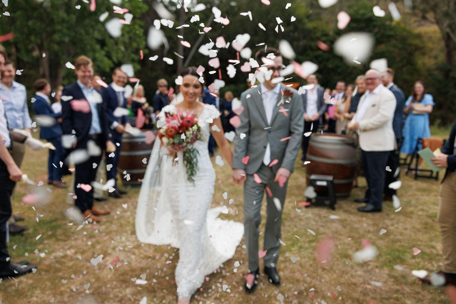 Bride and groom walk through confetti shower after outdoor wedding ceremony, guests celebrating around them.