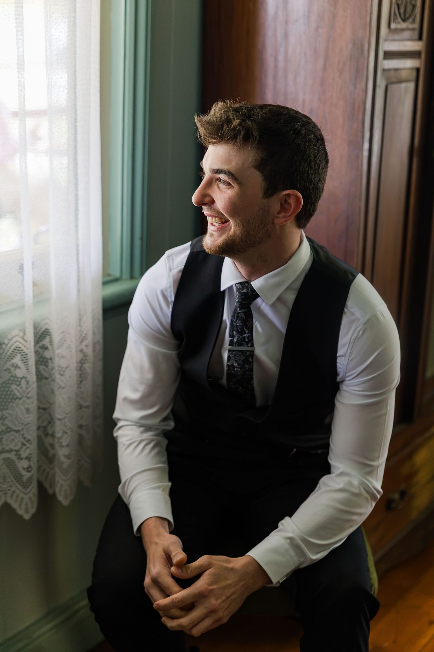 Smiling groom in white shirt, black vest, and tie sits by a window with lace curtains on his wedding day.