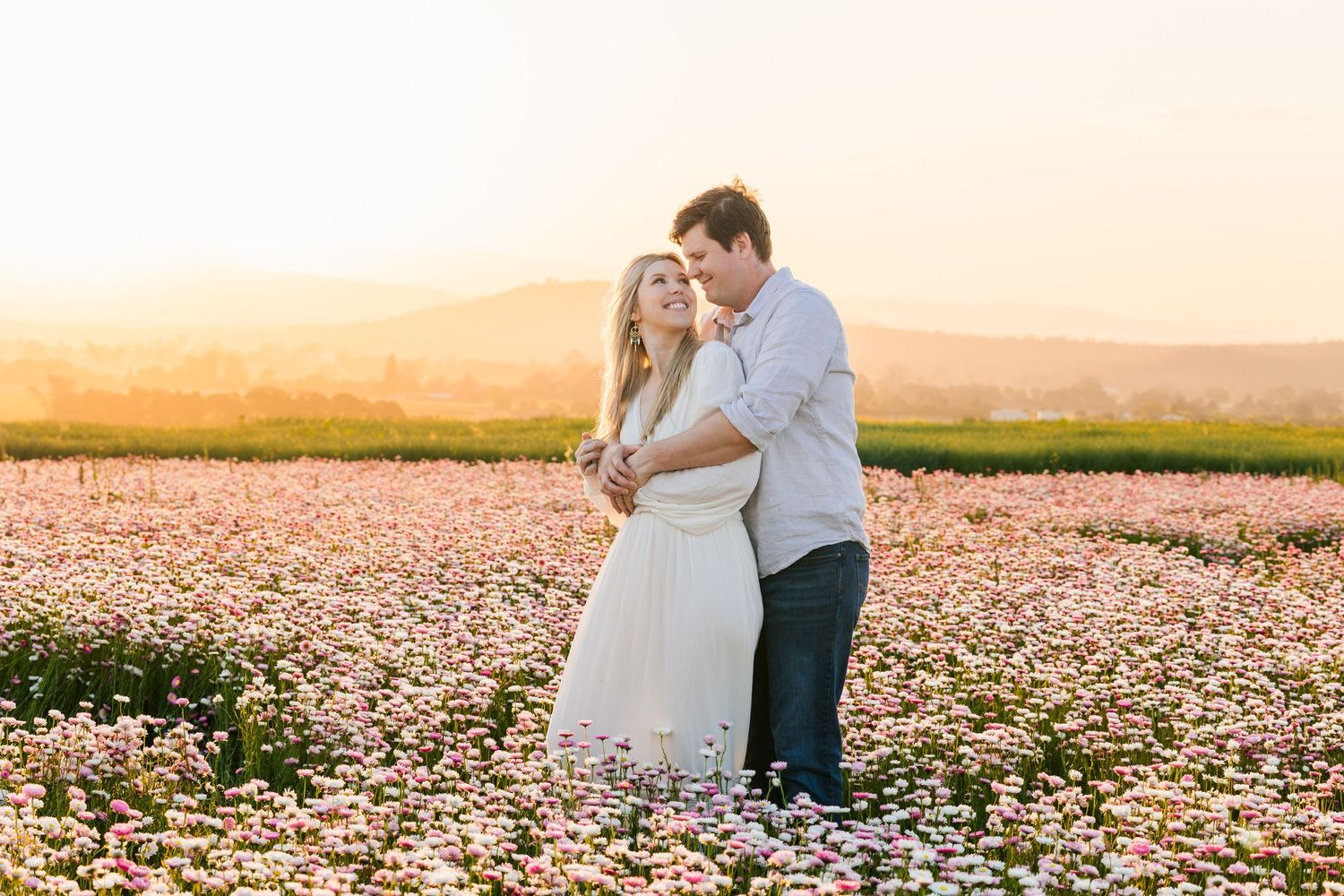 Couple embracing in a sunlit field of pink flowers with golden sunset and rolling hills in the background.