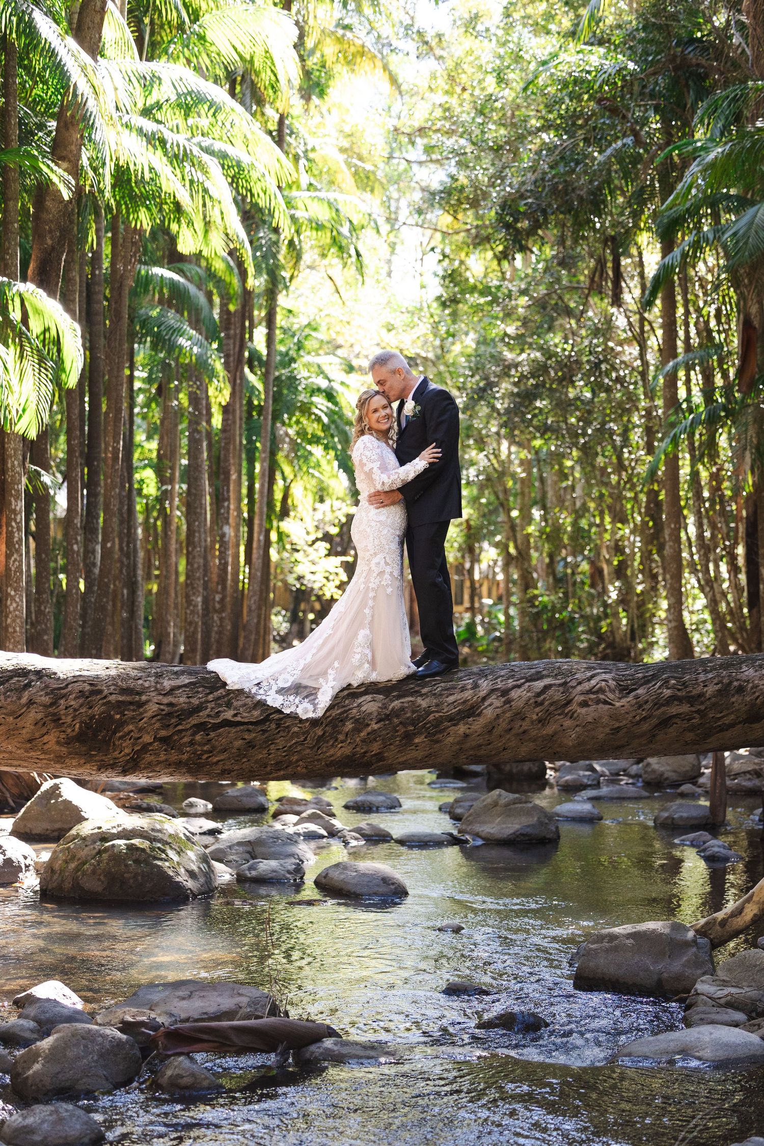 A couple shares an intimate moment on a fallen log over a tranquil stream in a lush tropical garden setting.