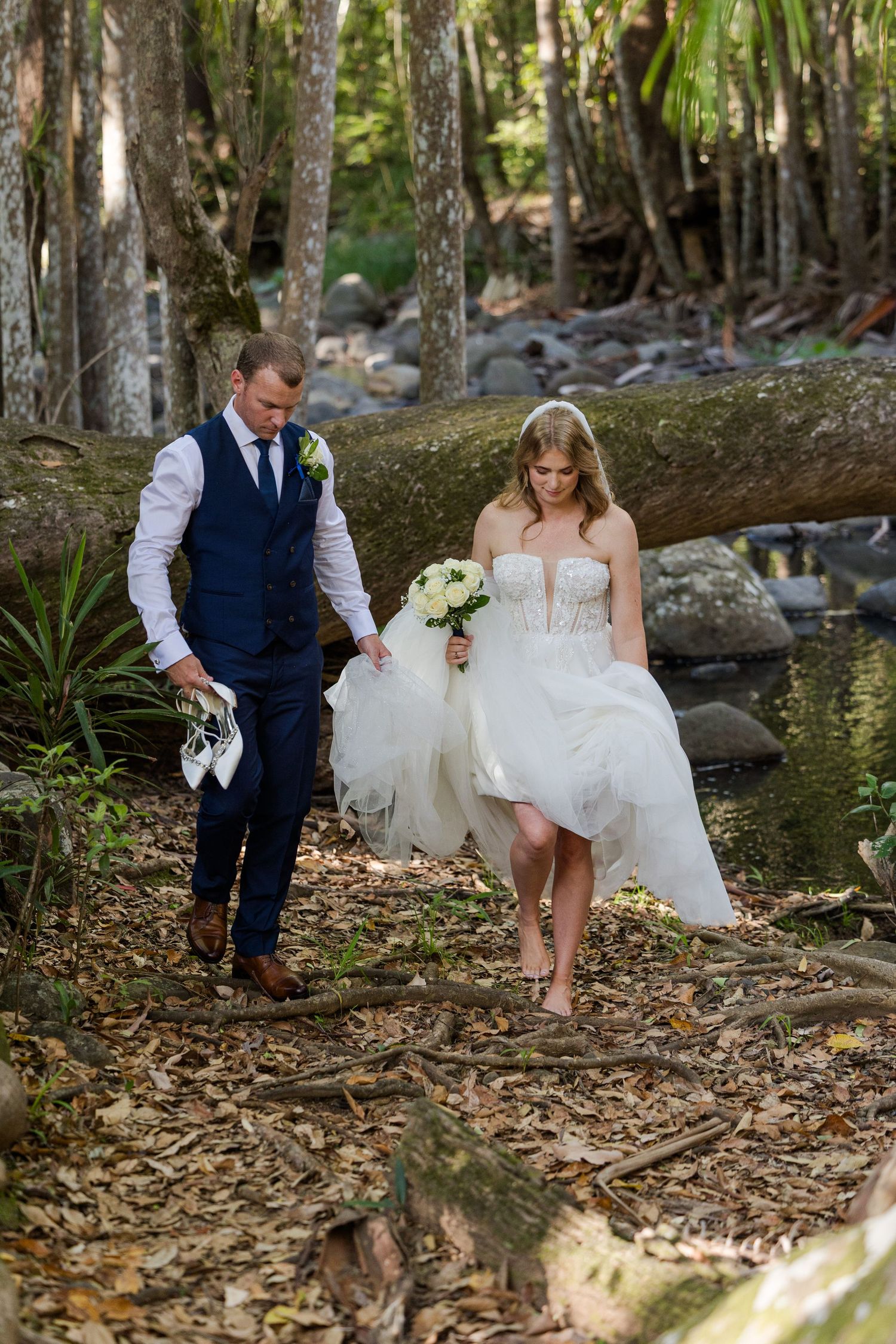 Wedding couple playfully walks through a lush forest path covered in fallen leaves.