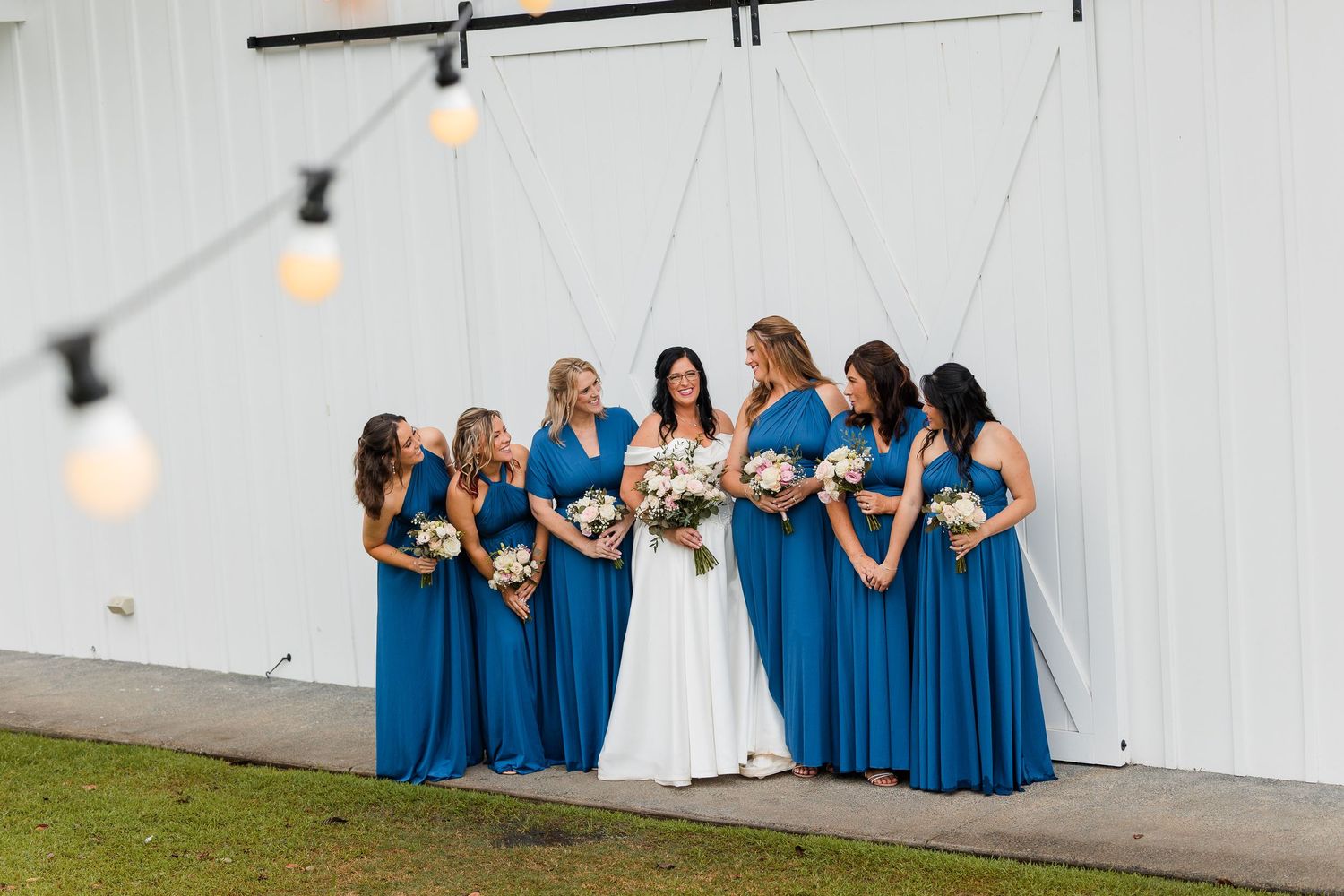 Bride in white gown with six bridesmaids in blue dresses holding bouquets outside a white barn wedding venue.