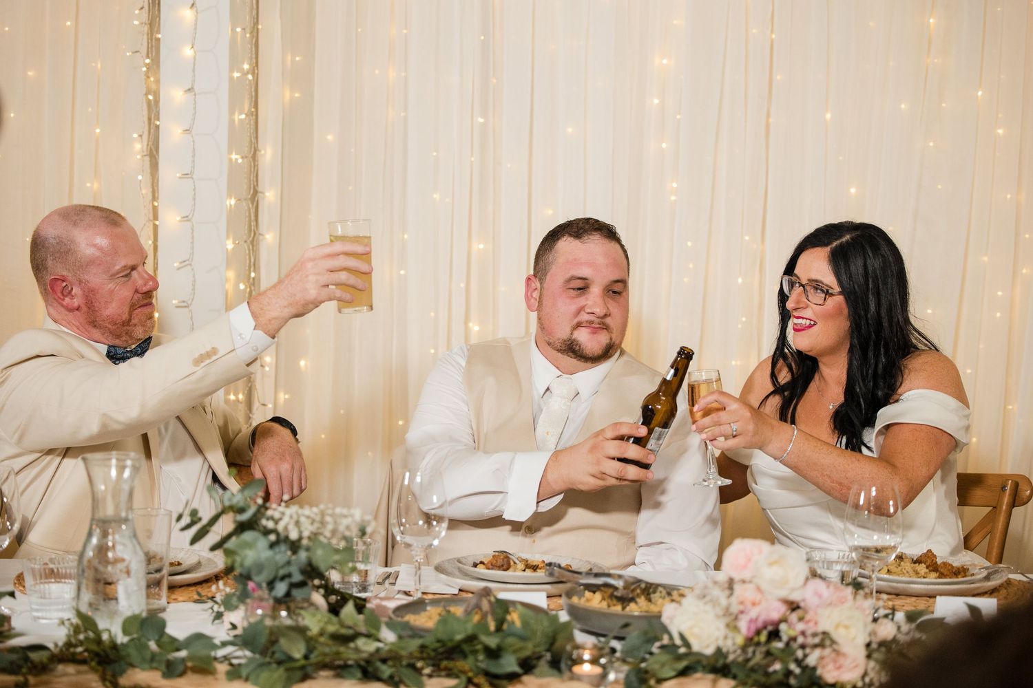 Bride and groom toasting with champagne at wedding reception table decorated with greenery and flowers.