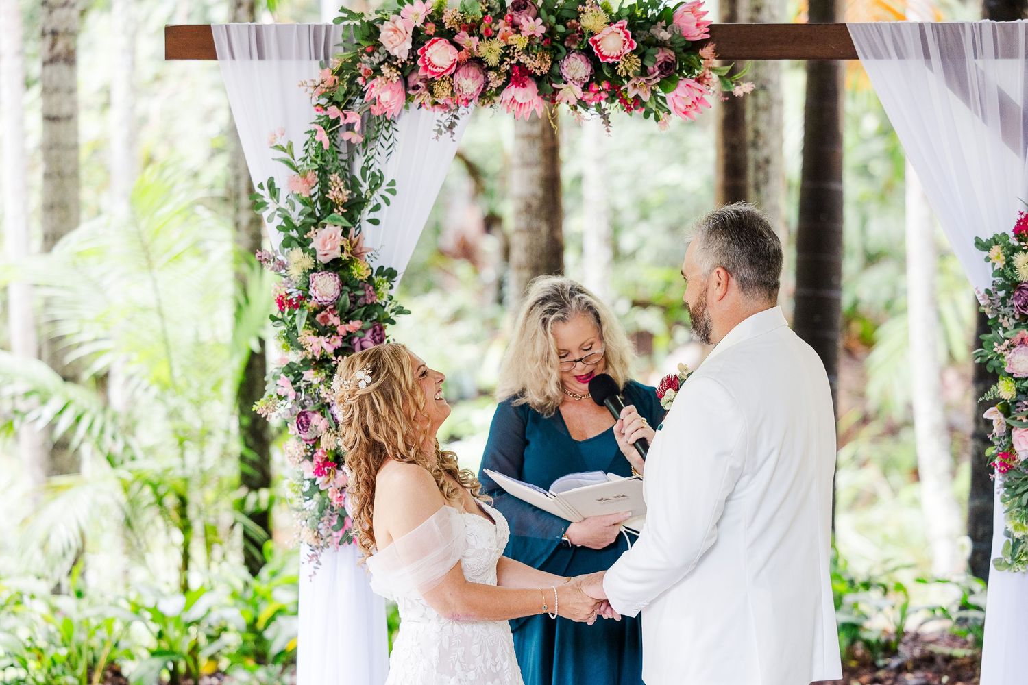 Couple exchanges vows under floral arch with pink roses, officiant in blue dress, tropical garden wedding ceremony.