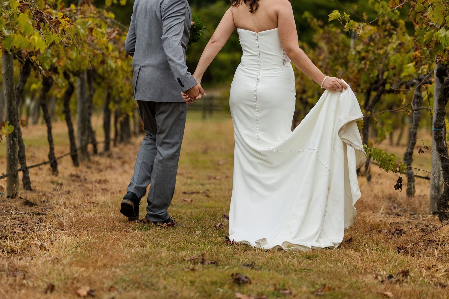 A bride and groom walk hand-in-hand through a vineyard, the bride in a white gown lifting her train.