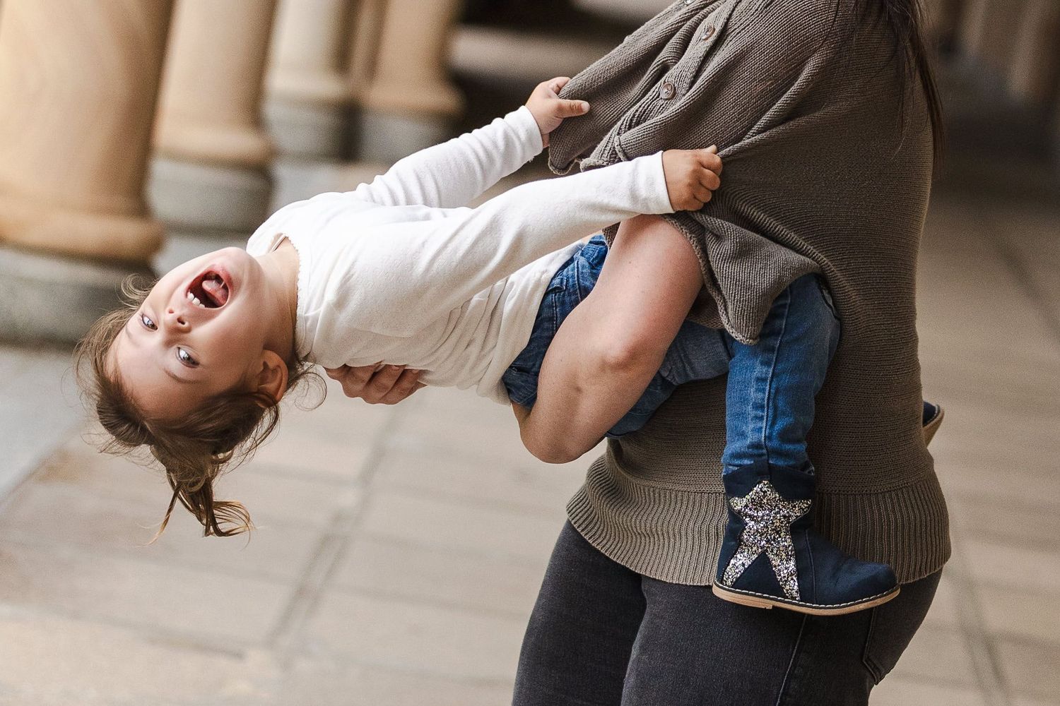 Person wearing a gray cardigan holds and playfully dips a laughing child in a white shirt and jeans.