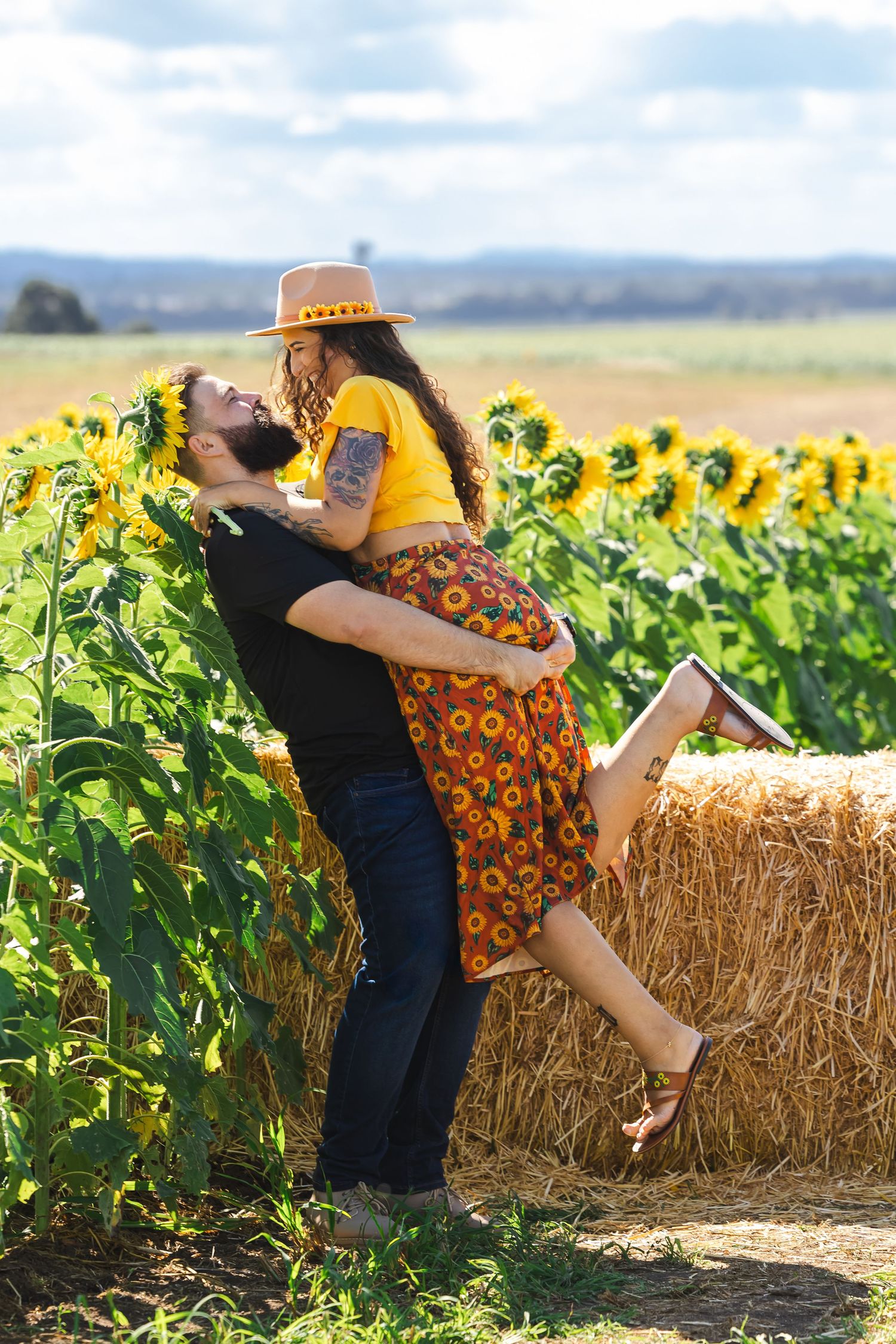 Romantic couple embracing and laughing near hay bales beside a sunflower field on sunny day.