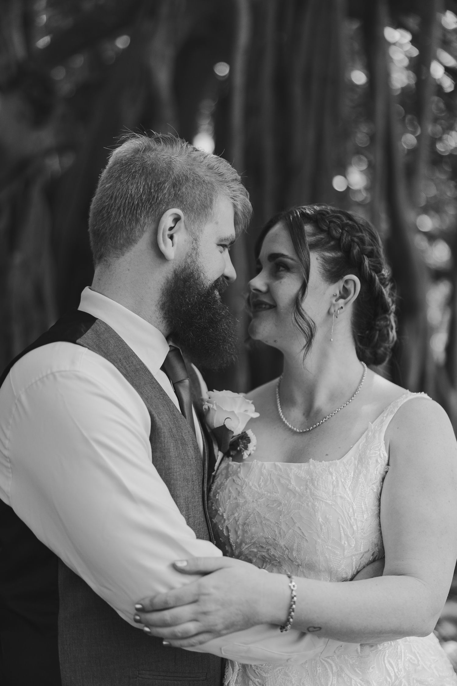 Black and white wedding photo of a bearded groom and smiling bride in lace dress embracing outdoors.