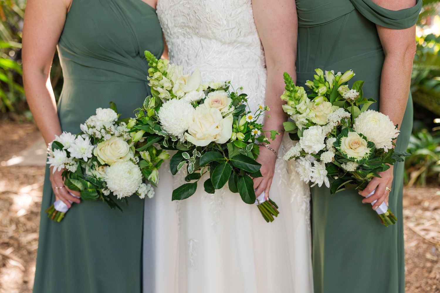 Bride and bridesmaids in sage green dresses holding white and green floral bouquets at a wedding ceremony.