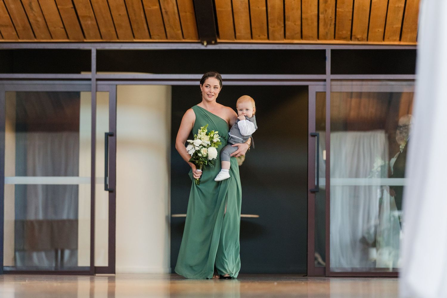 Bridesmaid in green gown holding baby and bouquet stands at wedding venue entrance.