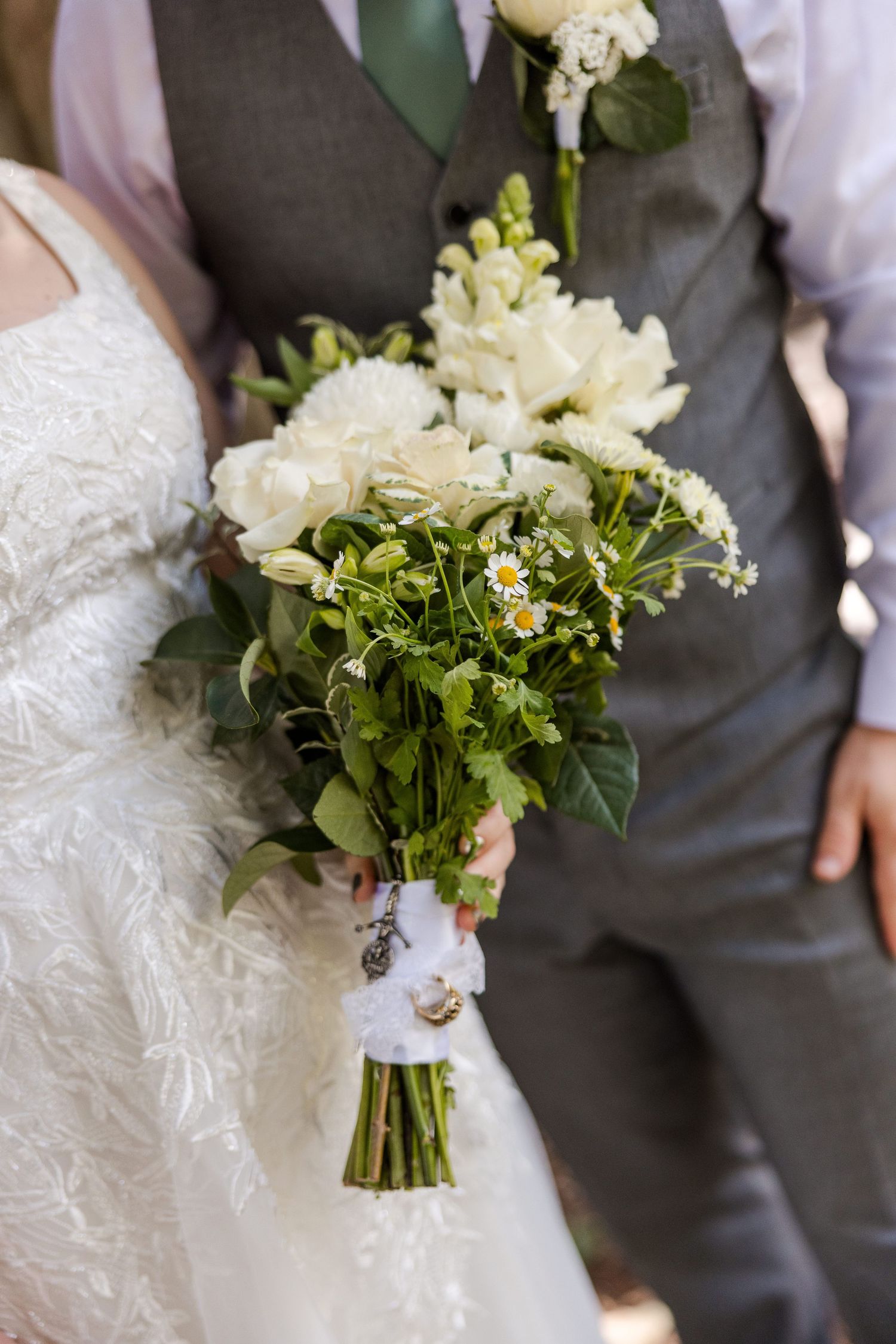 Bride holding a rustic white wedding bouquet with greenery and daisies, wrapped in white ribbon, standing beside groom in gray vest.