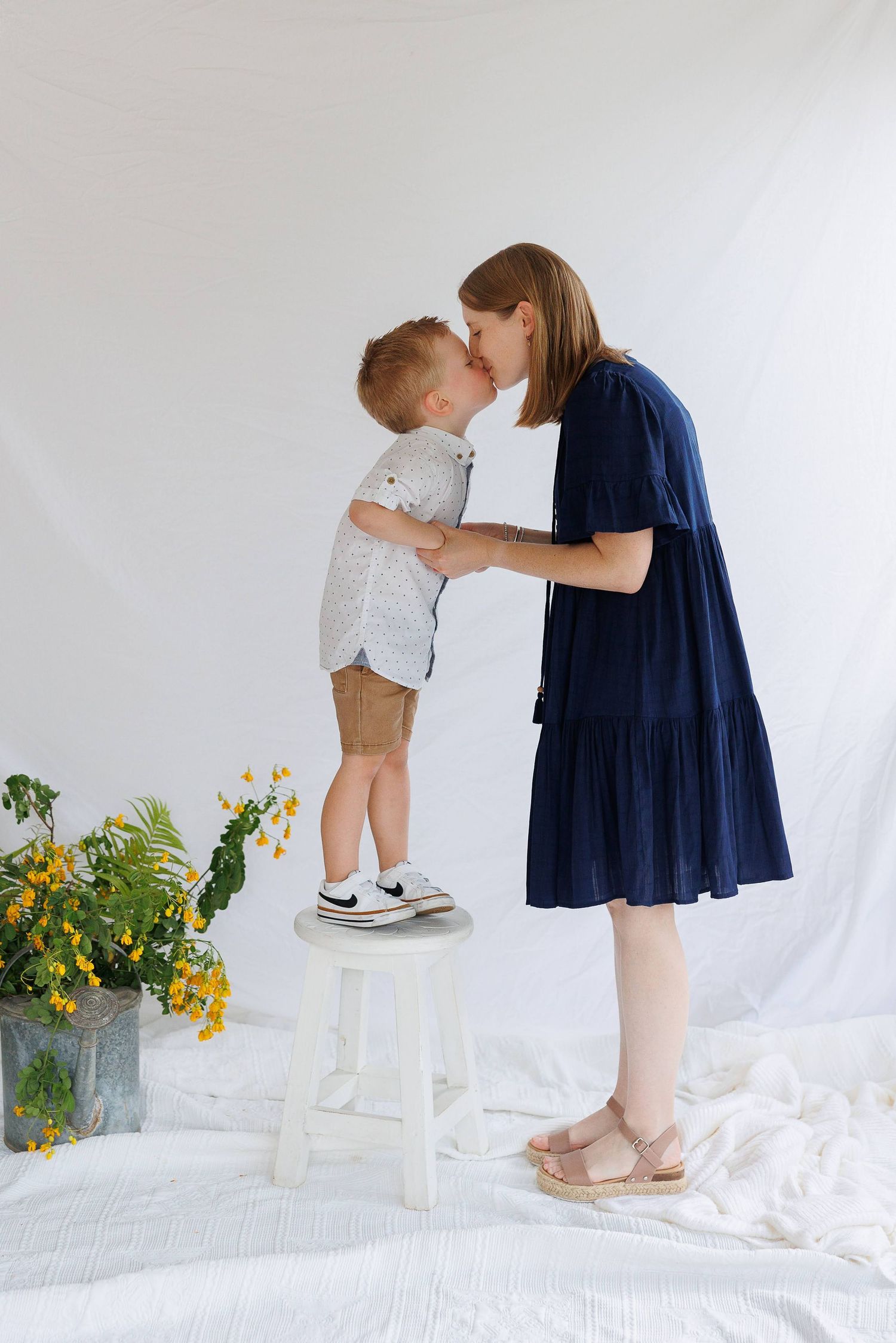 A mother in a navy dress kisses her toddler son standing on a white stool in a bright studio setting.