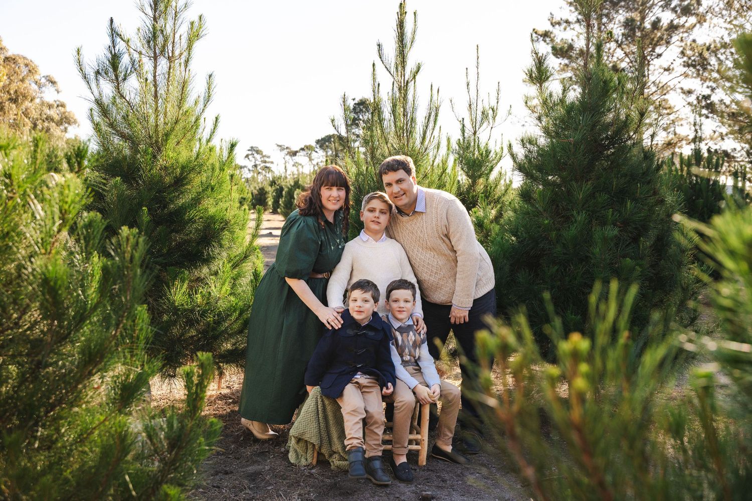 Family sits together on wooden bench surrounded by evergreen trees during outdoor portrait session.
