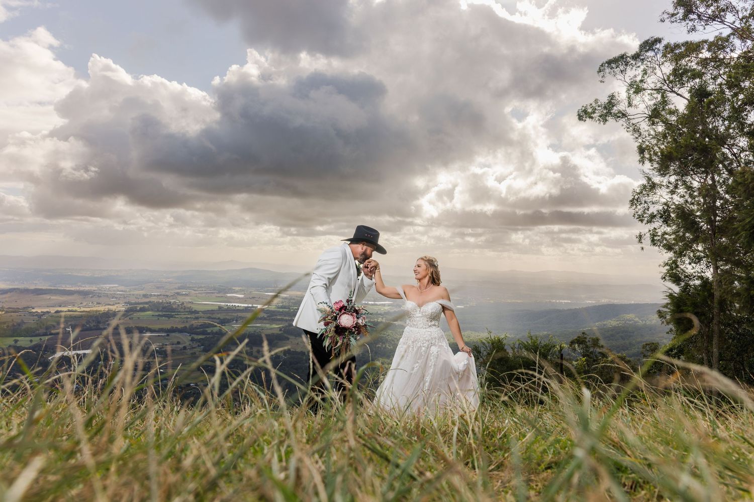 Bride and groom holding hands on a hilltop with dramatic cloudy skies and scenic valley views on their wedding day.