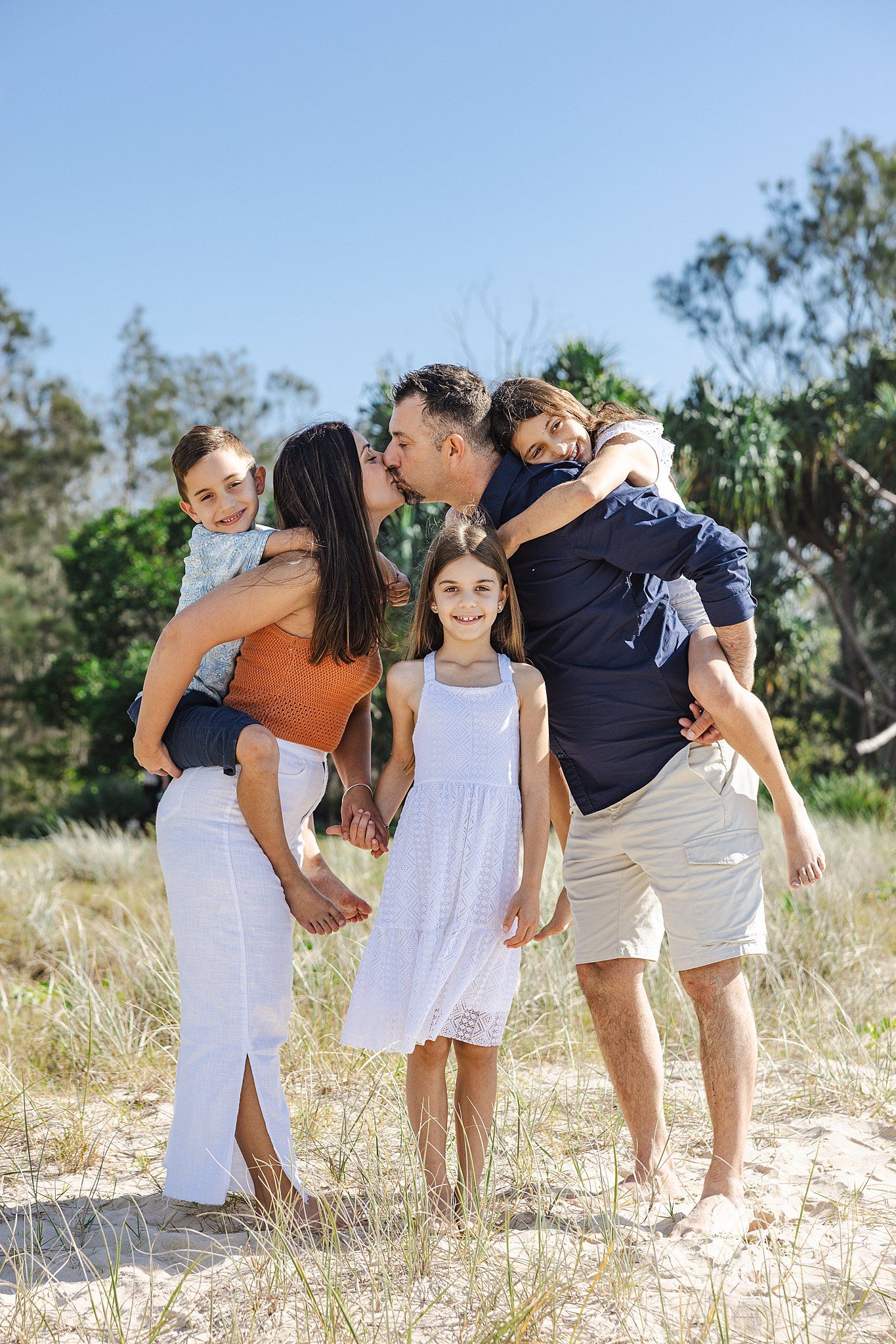 A family of four standing together outdoors in sand dunes with green foliage on a sunny day.