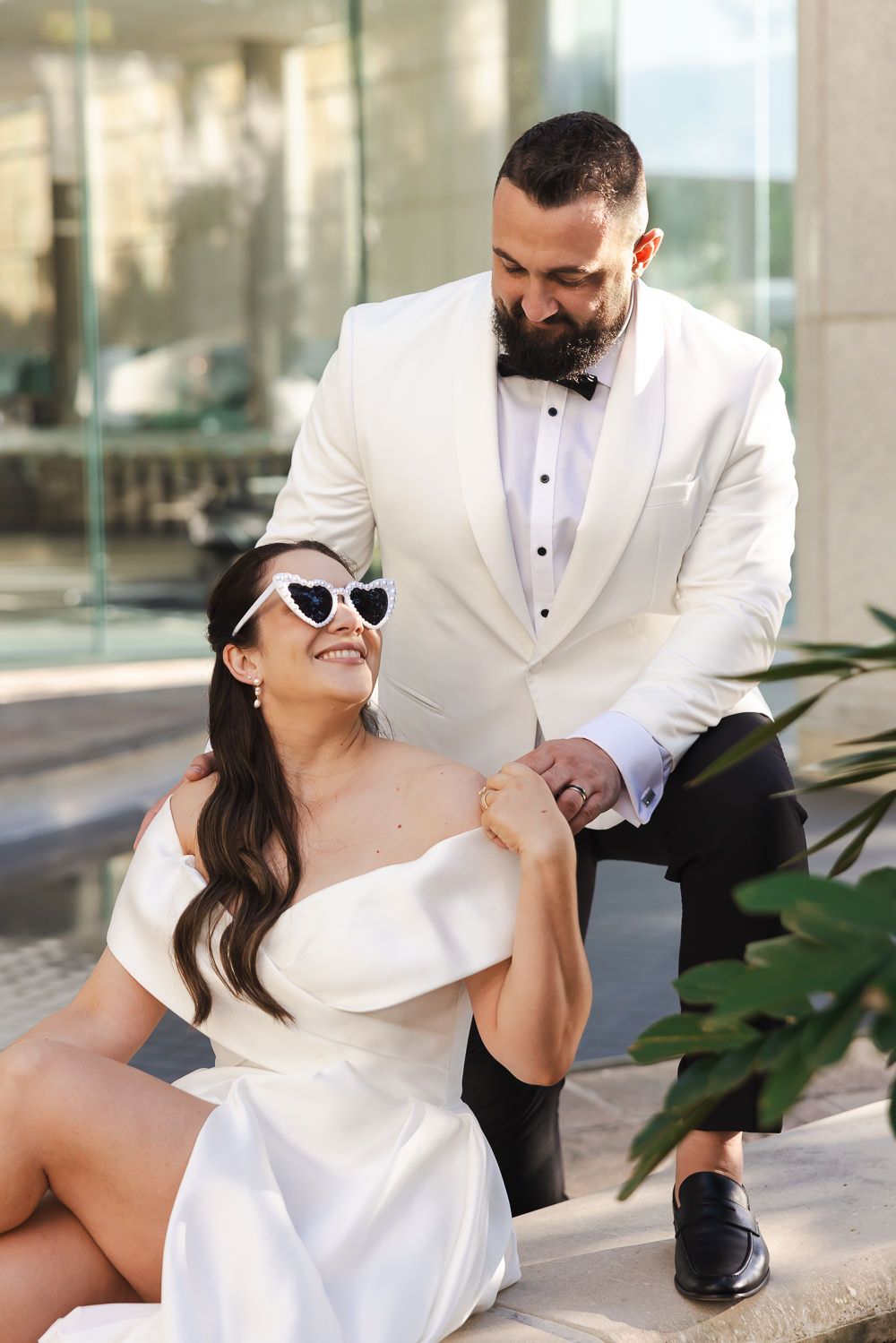 A couple in white formal attire poses together outdoors, with sunglasses and fashion-forward styling.