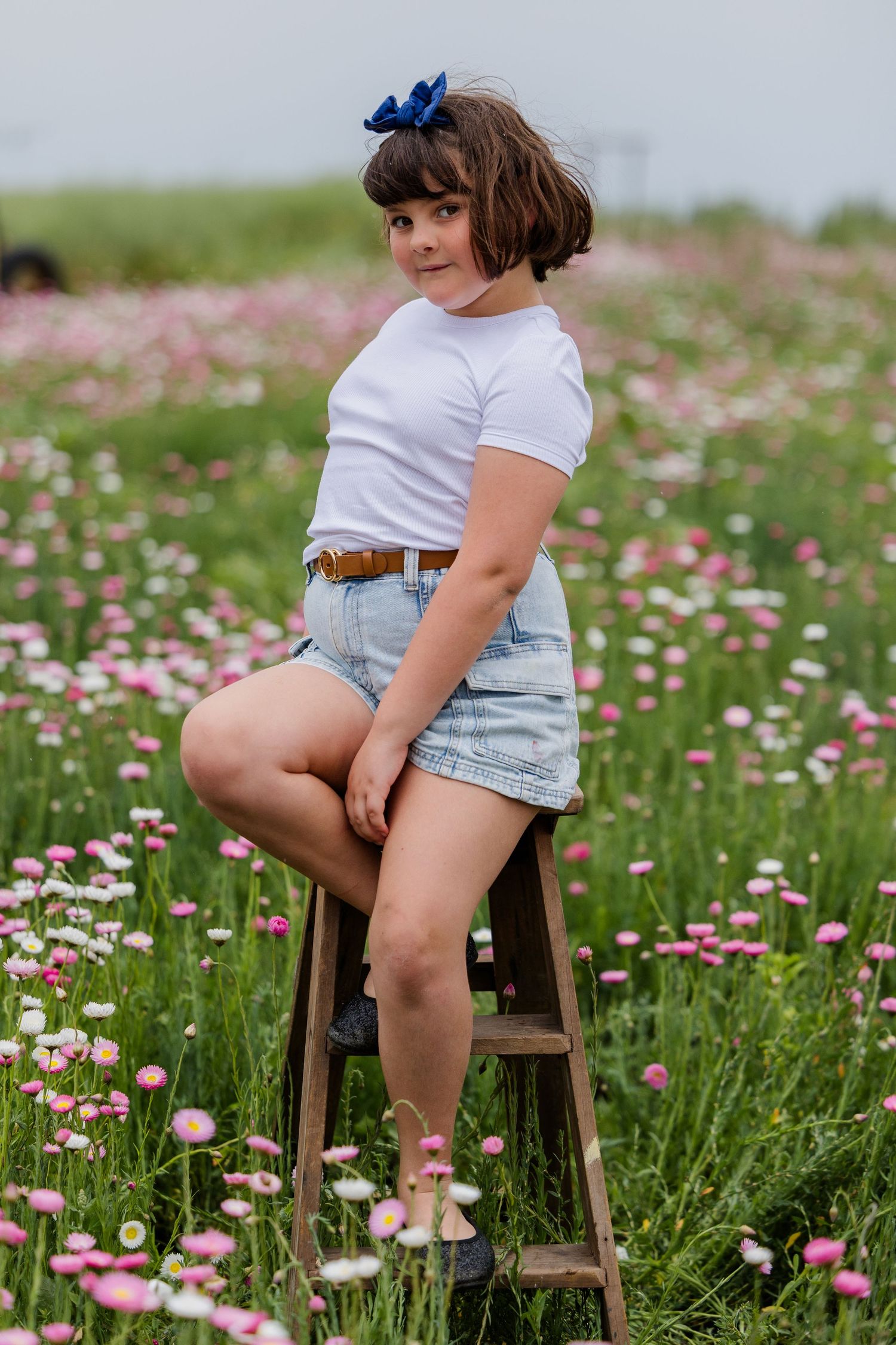Young woman in white tee and denim shorts poses on wooden ladder in a vibrant pink wildflower field.