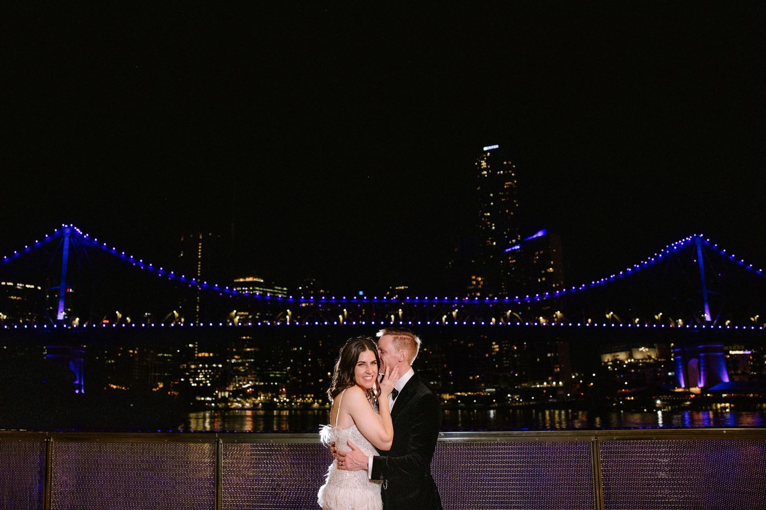 A couple dances romantically at night with the illuminated Benjamin Franklin Bridge glowing blue in Philadelphia's skyline.