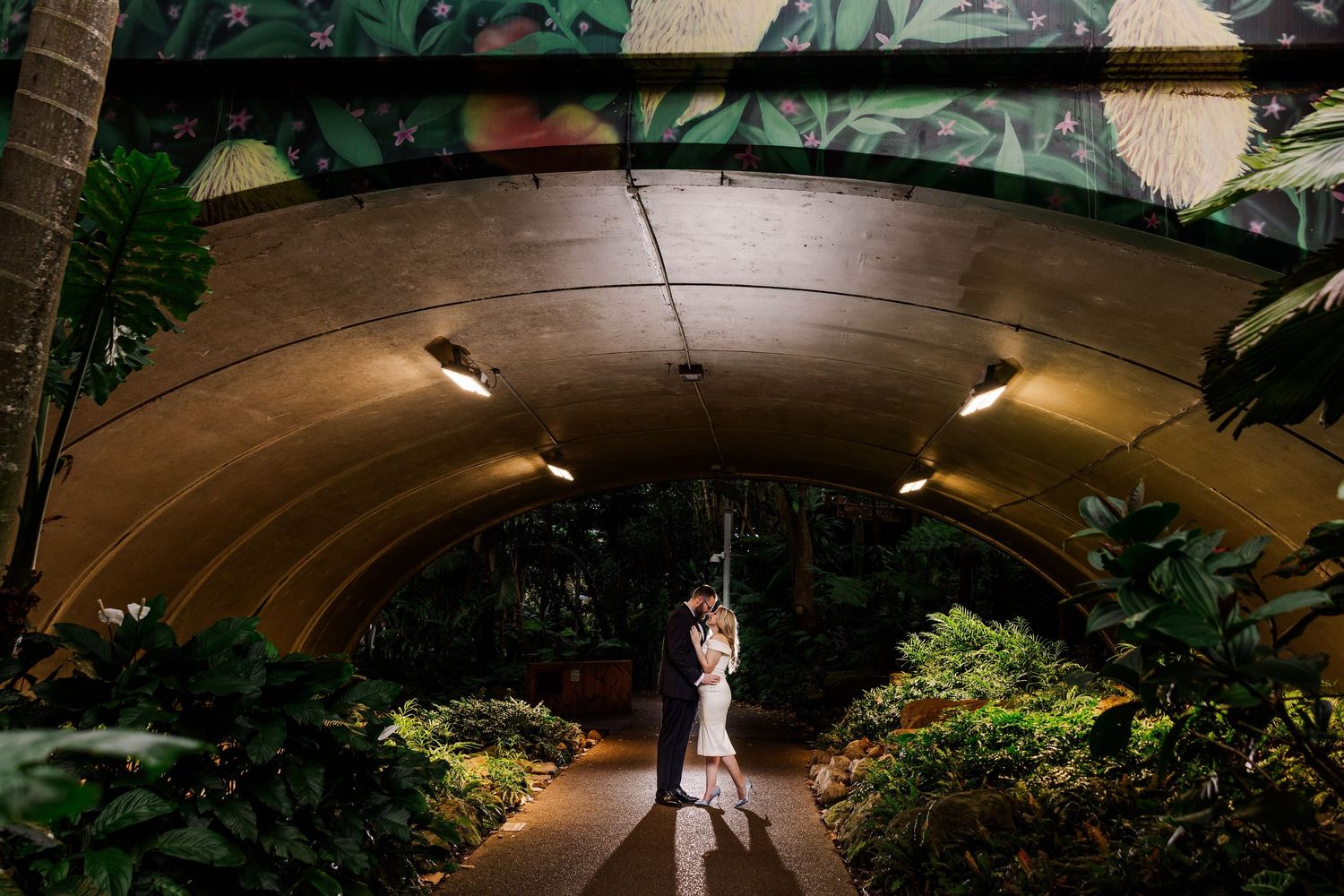 Illuminated curved tunnel pathway surrounded by lush green plants and modern lighting fixtures creates a futuristic atmosphere.