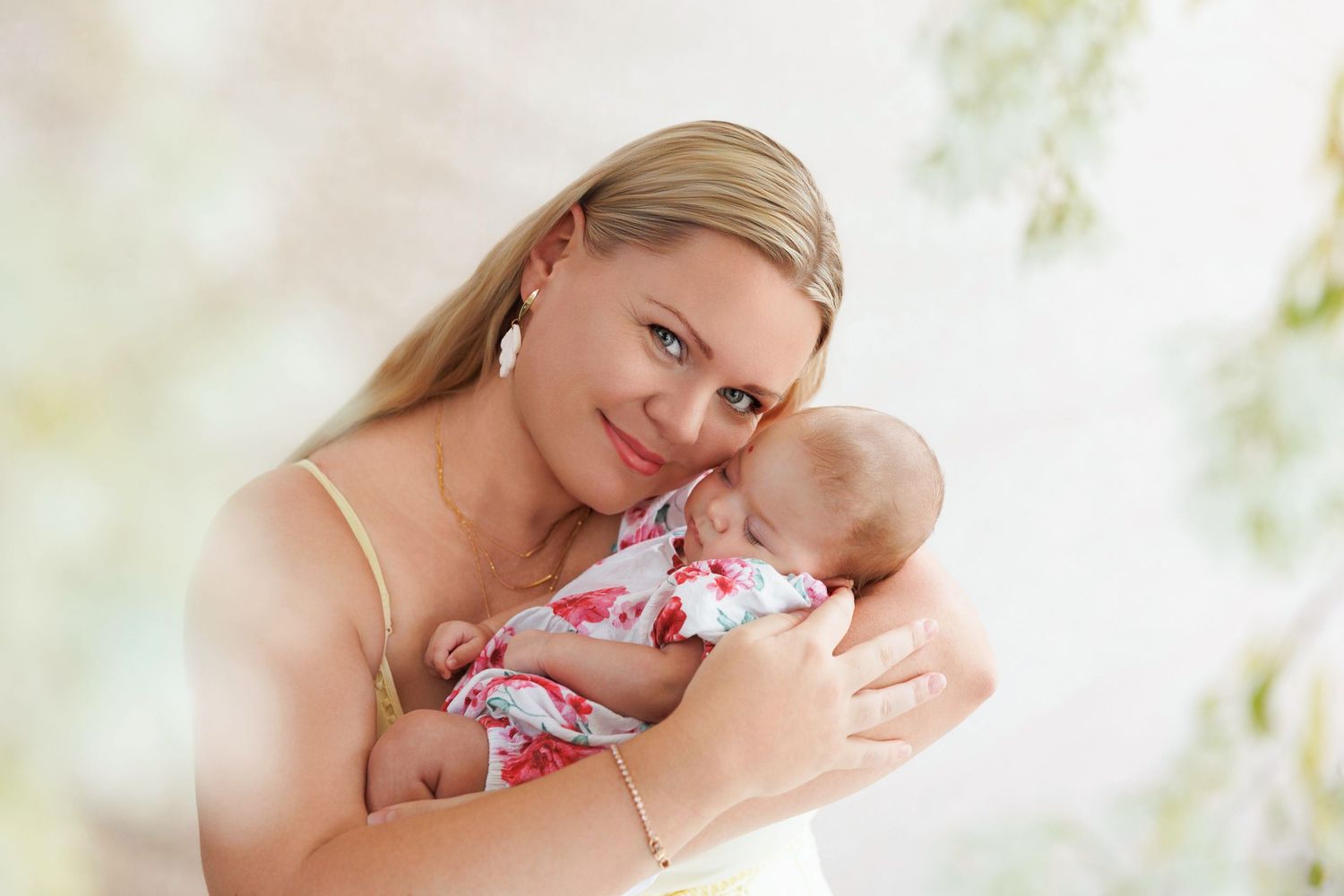 A young blonde mother tenderly holds her sleeping newborn baby dressed in a floral outfit against a soft pastel background.