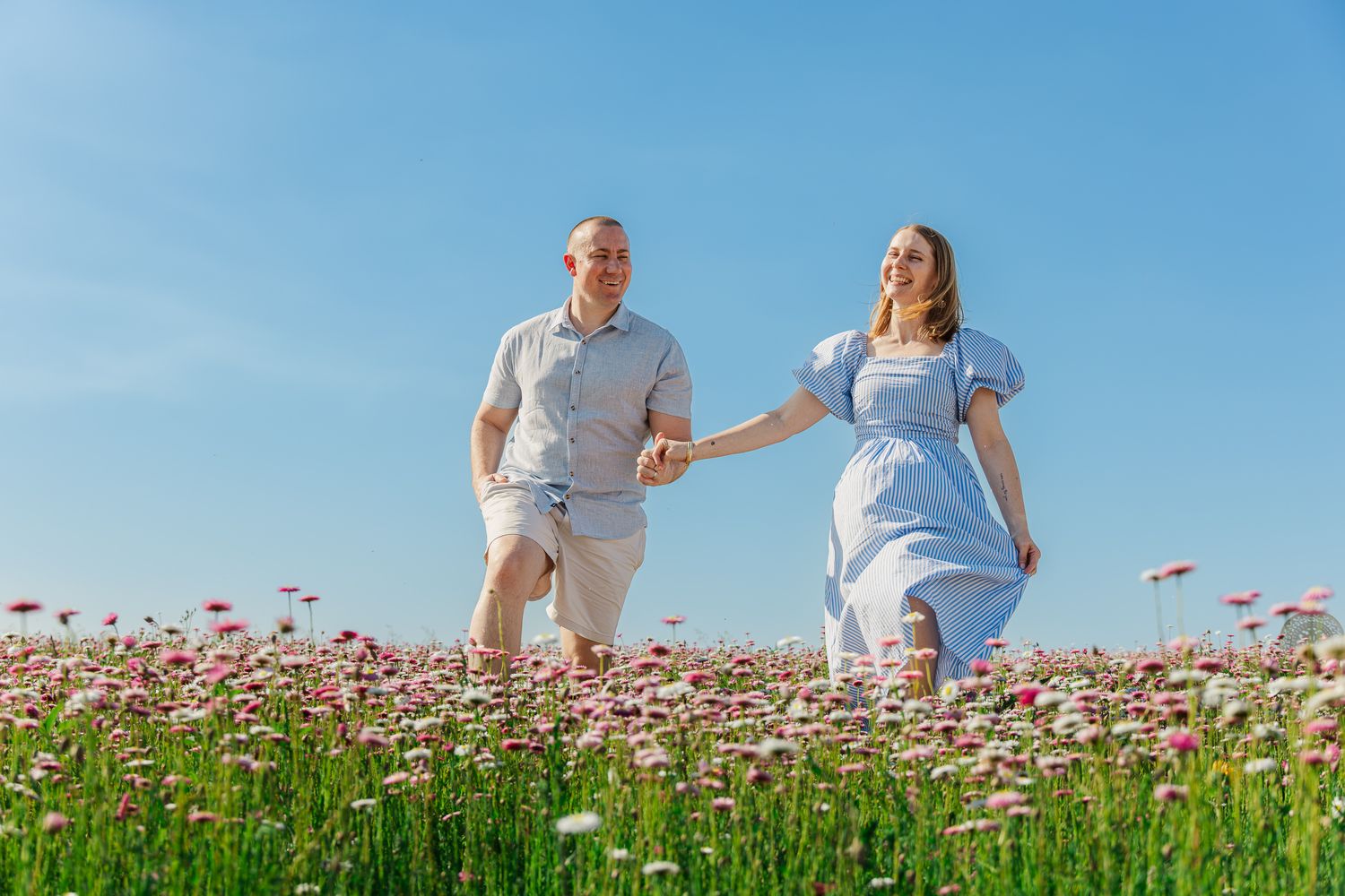Couple holding hands running through a blooming flower field under a clear blue sky on a sunny day.