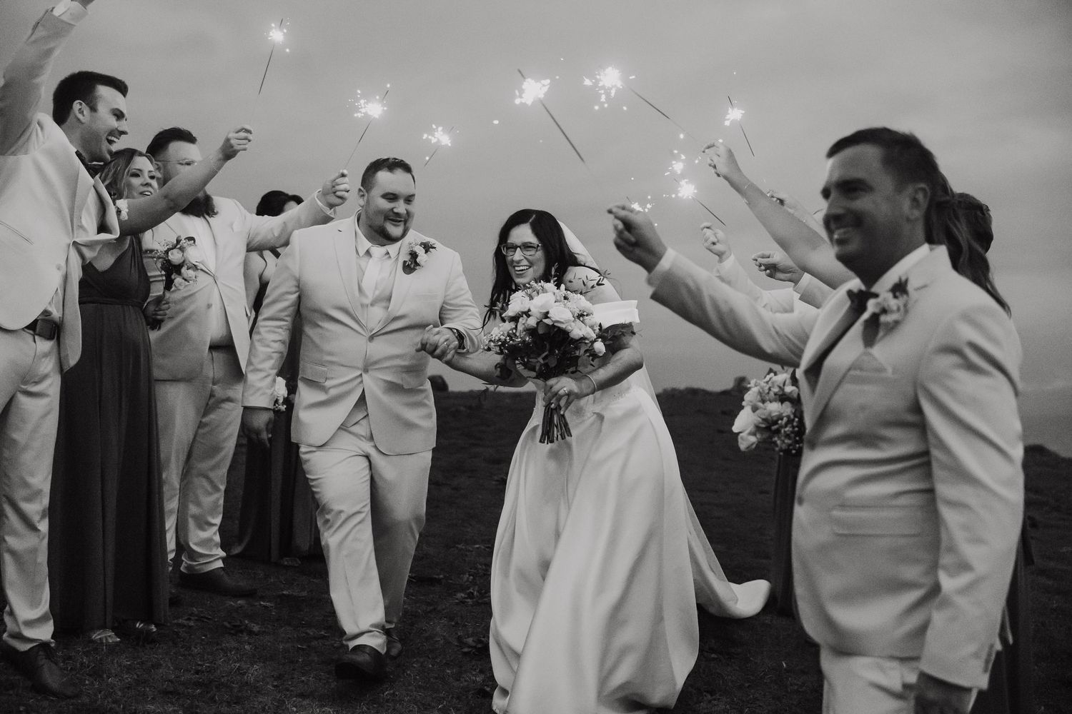 Black and white photo of bride and groom walking through sparkler send-off with wedding party cheering.