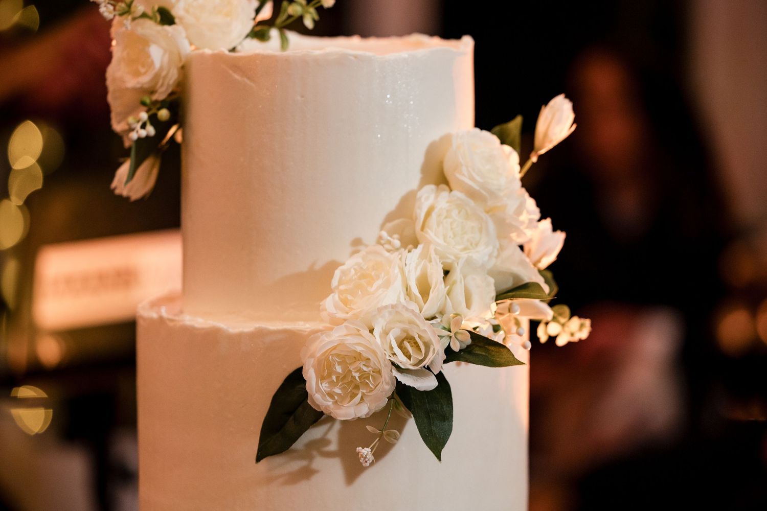 Elegant white two-tier wedding cake adorned with fresh white roses and green leaves on a dark reception background.