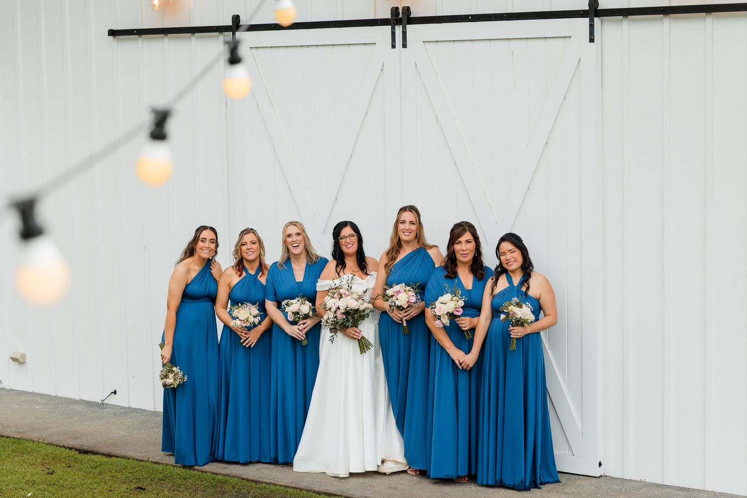 Bride in white gown with six bridesmaids in blue dresses holding bouquets in front of white barn doors.