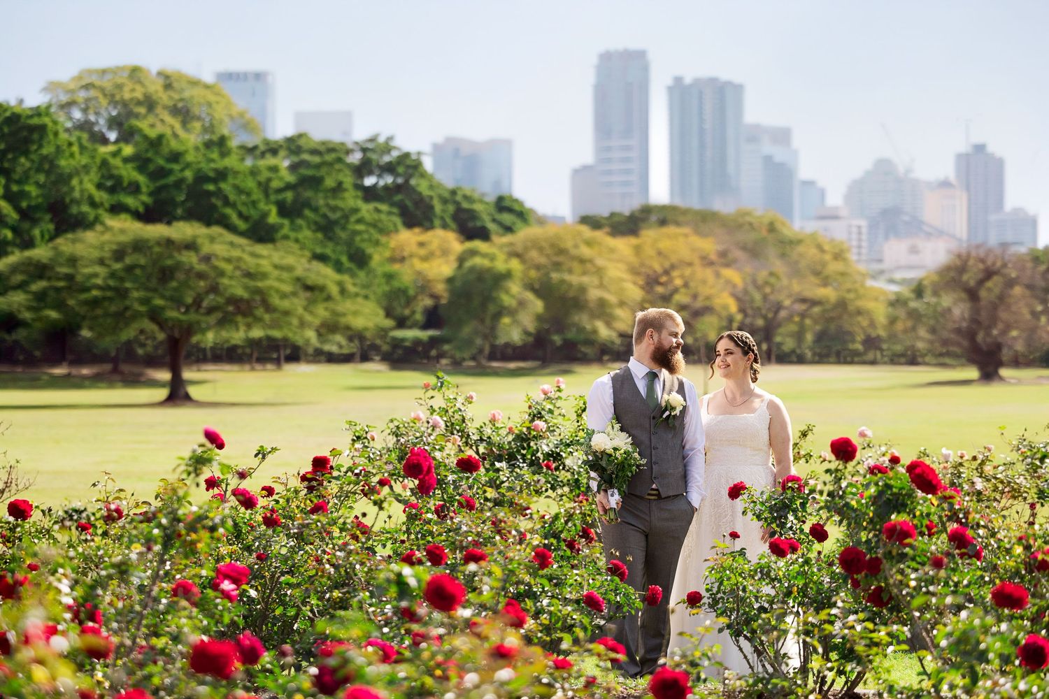 Bride and groom standing among red roses in a lush green park with city skyline in the background.