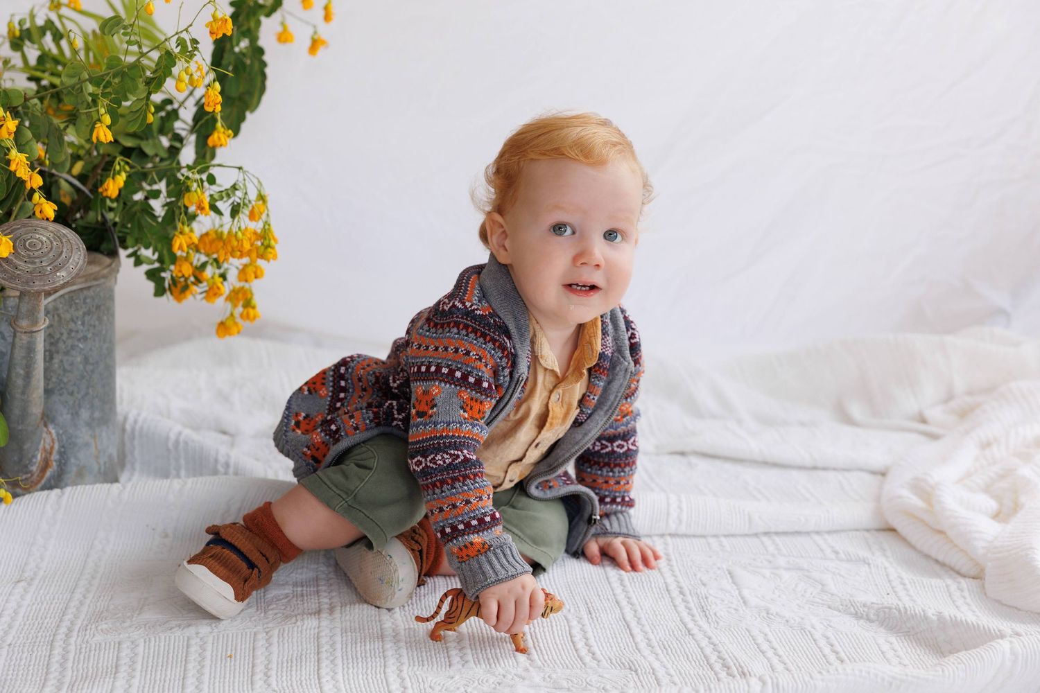 Adorable baby boy in a colorful patterned cardigan and green pants sitting on a white blanket near yellow flowers.