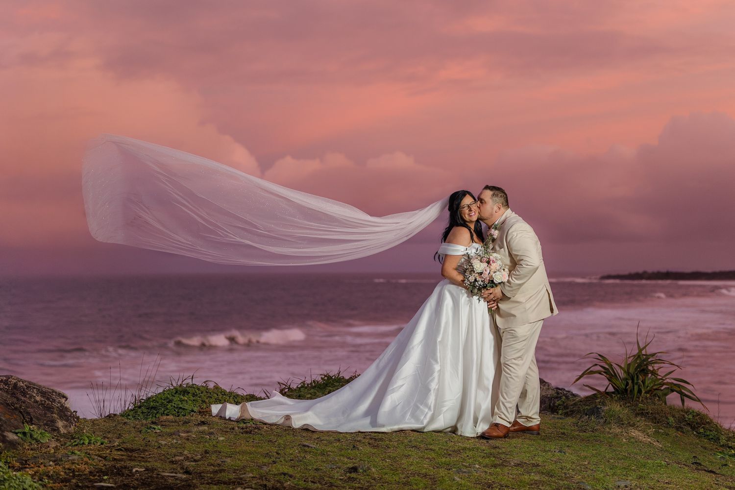 Bride and groom share a romantic kiss at sunset by the ocean, with veil flowing in the wind against a pink sky.