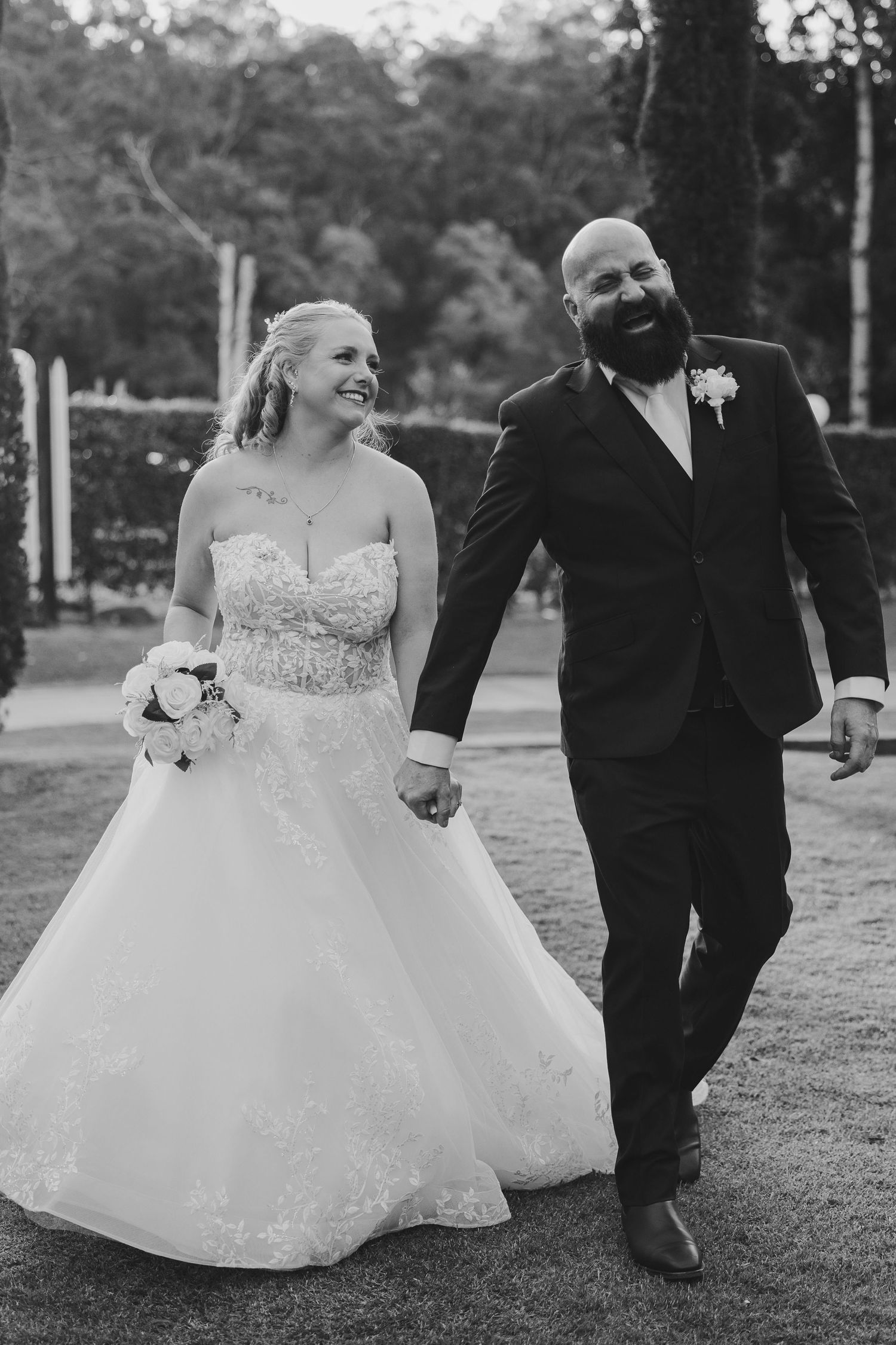 Black and white photo of a smiling bride and groom walking hand in hand outdoors on their wedding day.