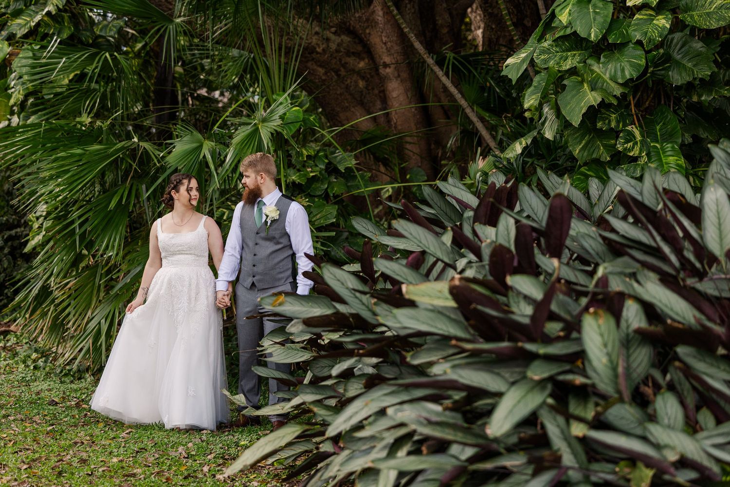 Bride and groom holding hands in a lush tropical garden surrounded by palm trees and colorful foliage on their wedding day.