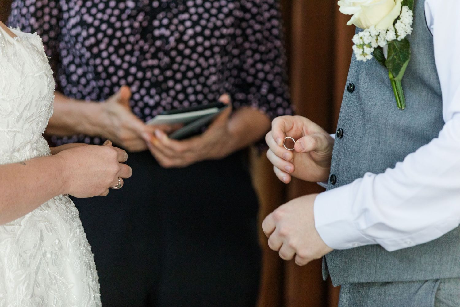 Groom placing wedding ring on bride's finger during ceremony, officiant holding phone in background.