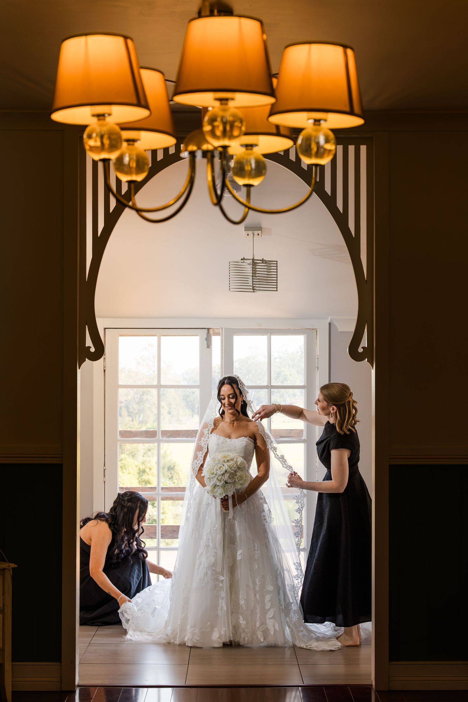Bride in white gown holds bouquet while bridesmaids assist her, framed by elegant arch with chandelier above.