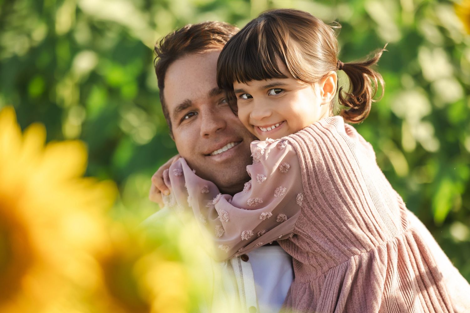 Smiling father and daughter hugging outdoors in a vibrant sunflower field on a sunny day.