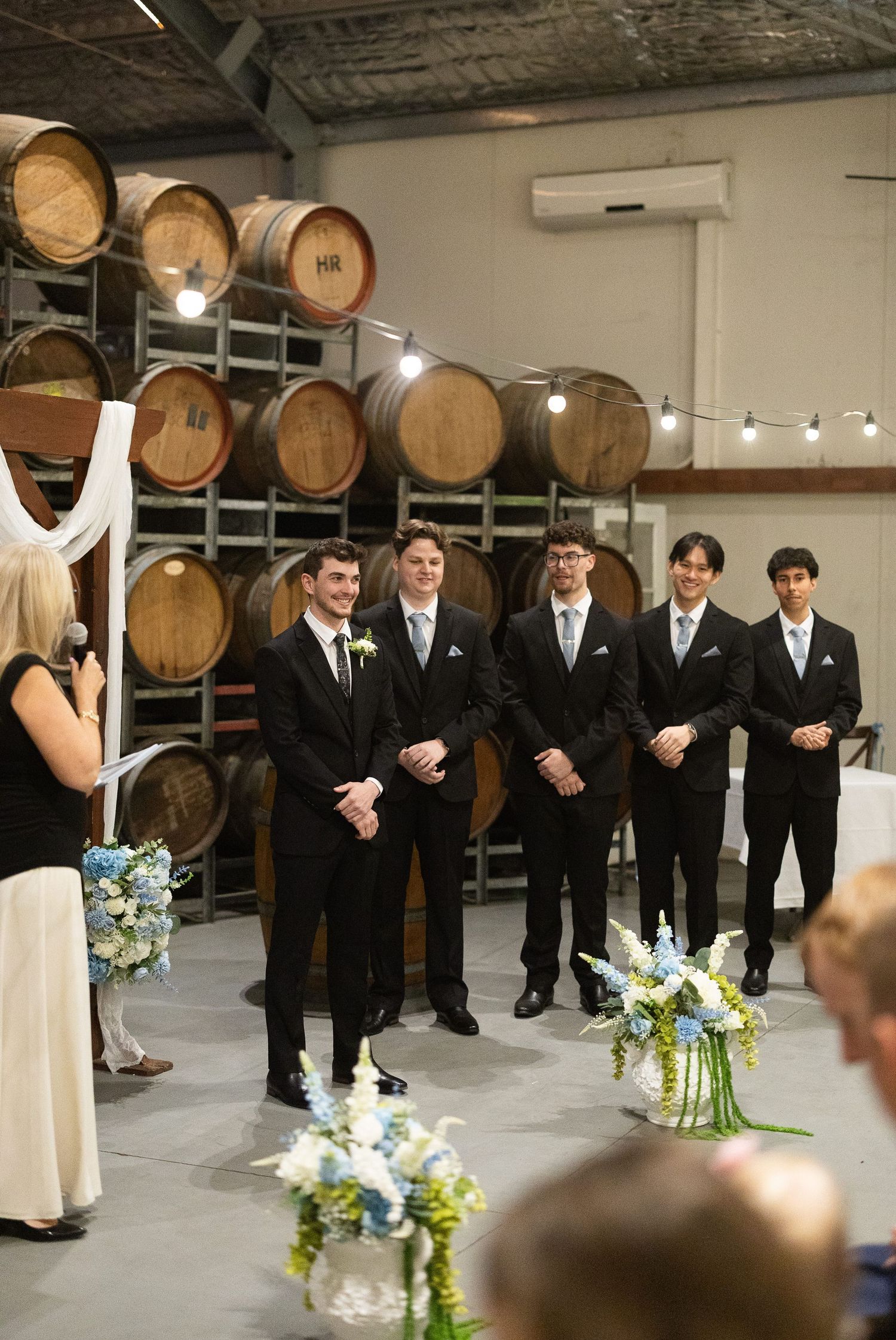 Groomsmen in black suits stand at winery wedding altar surrounded by oak barrels and blue floral arrangements.