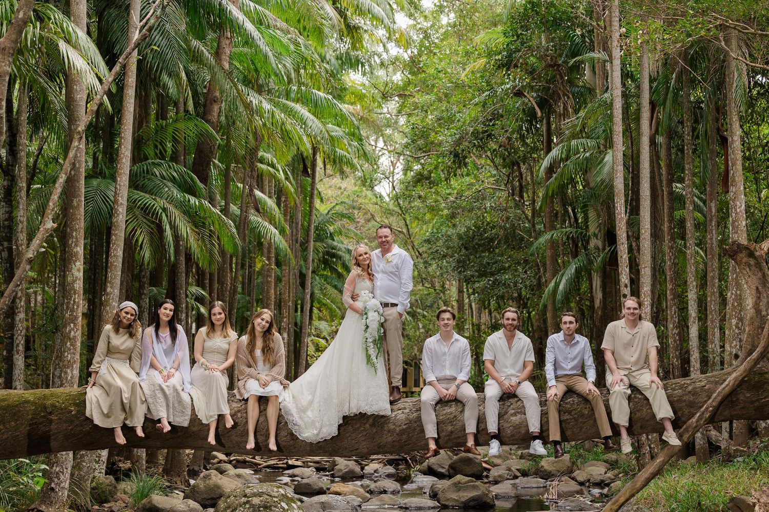 Wedding party poses on a rustic bridge surrounded by lush tropical forest, bride in white gown with groomsmen and bridesmaids.