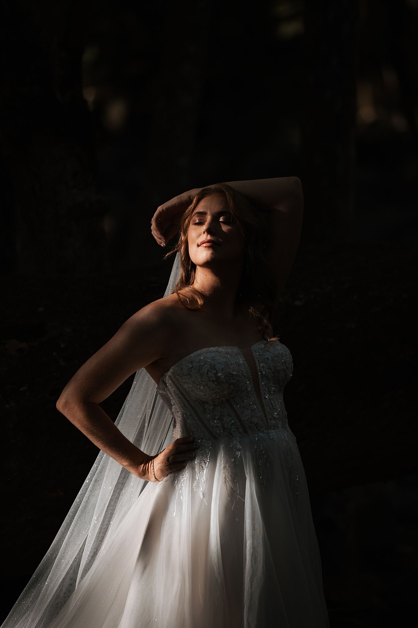 A figure in a flowing white wedding gown poses dramatically in moody lighting against a dark background.
