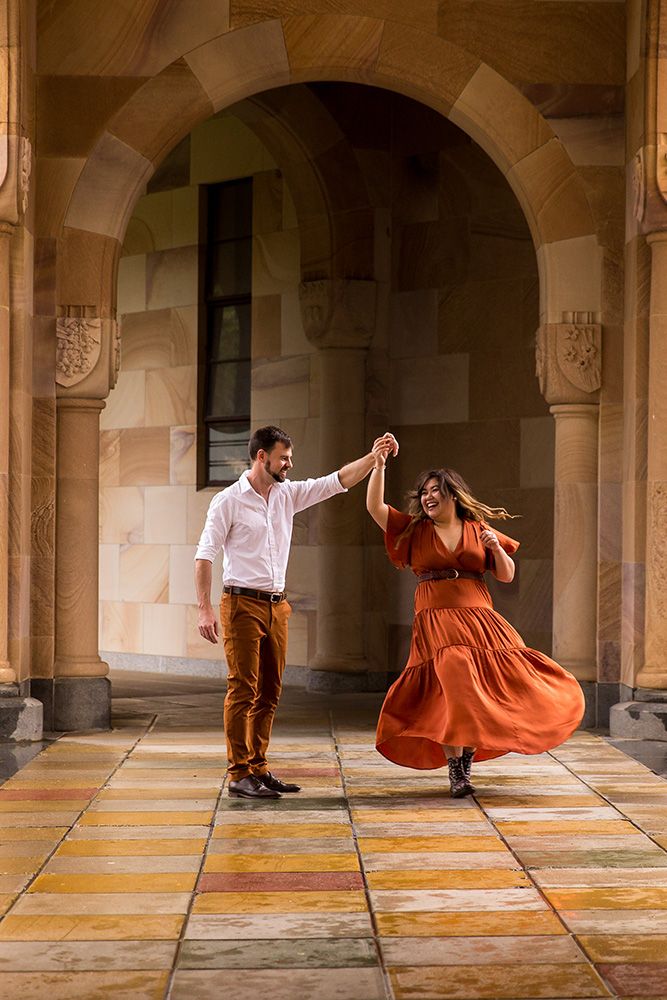 Couple dancing playfully under stone archway with woman in flowing orange dress.
