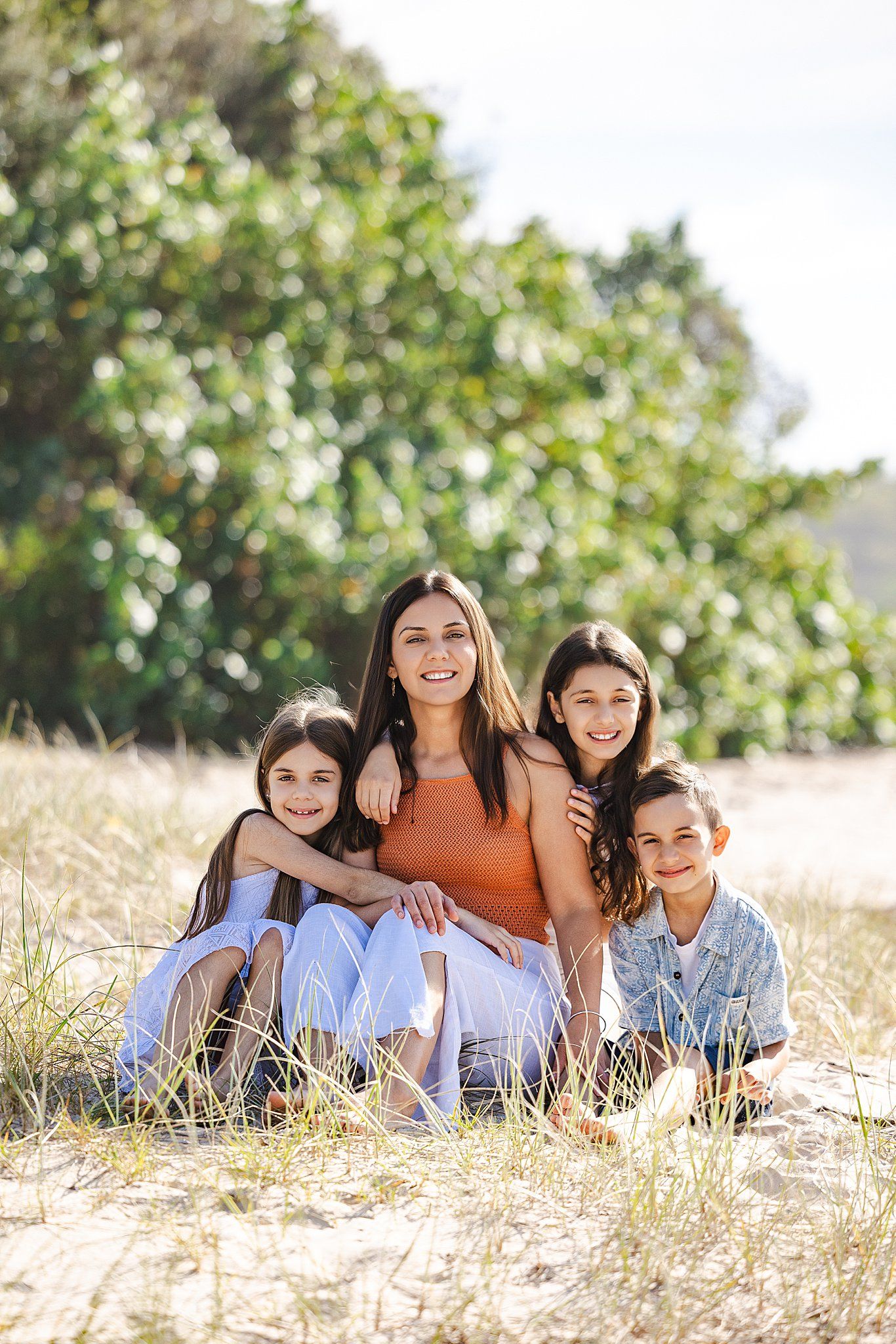 Family sitting together on sandy ground with green foliage in background on sunny day.