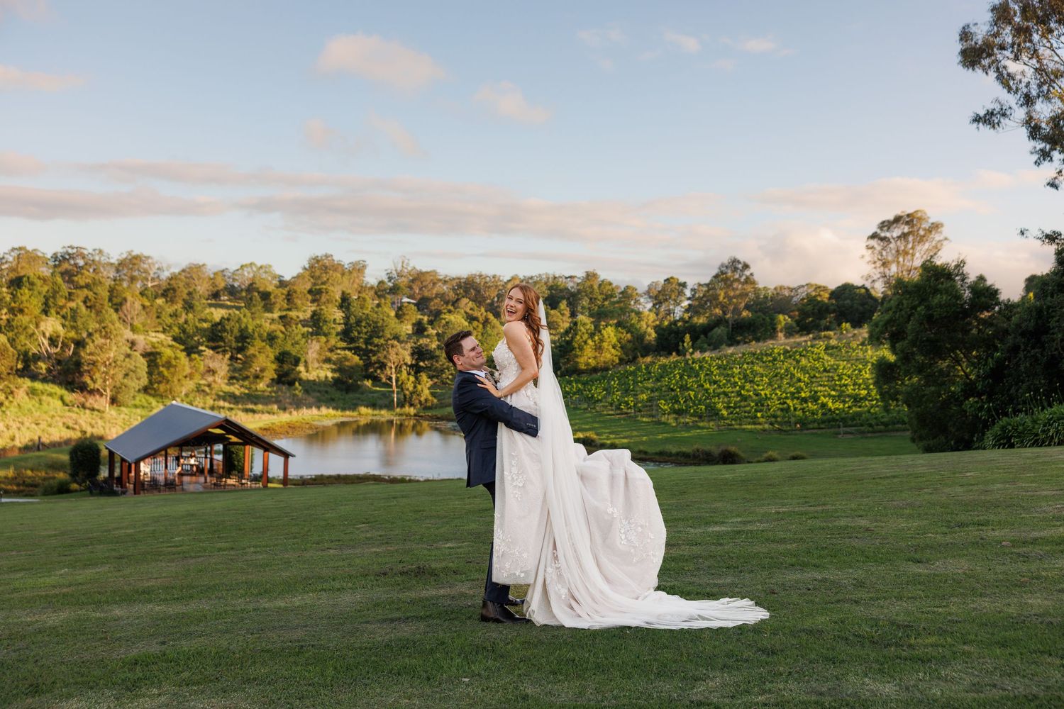 Couple in wedding attire stands on a hilltop lawn overlooking a lake and vineyard during golden hour.