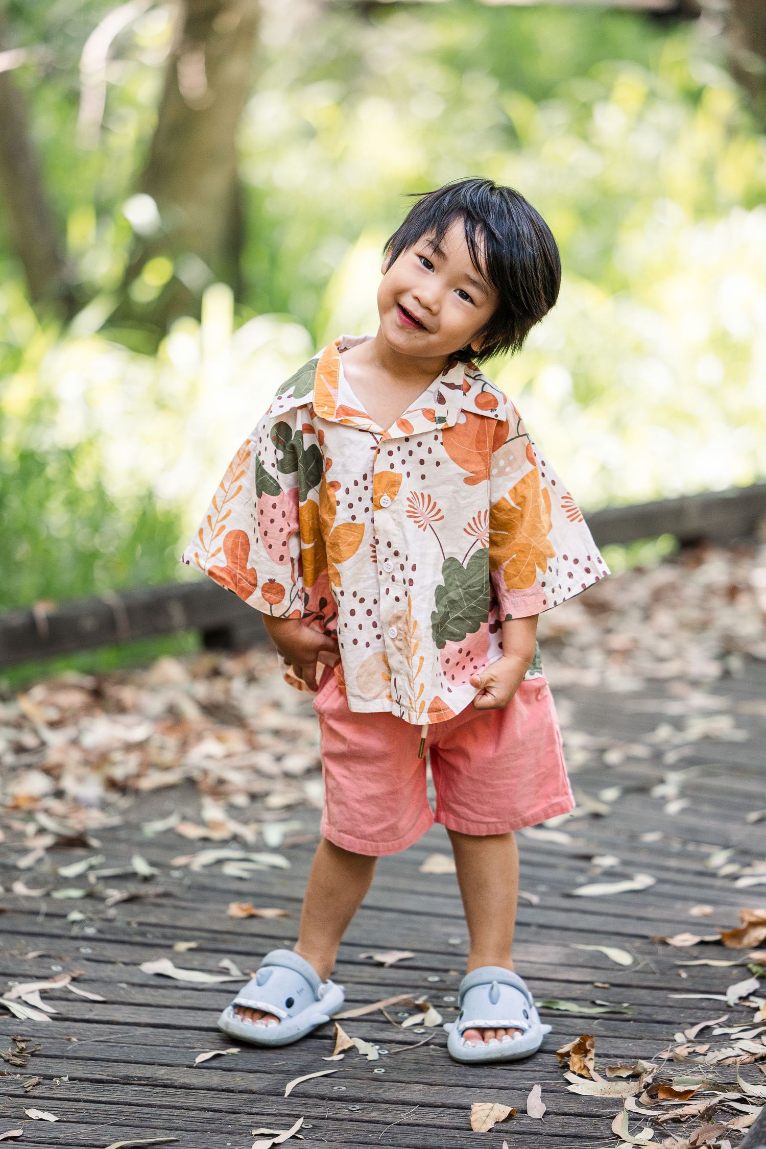 A child in a floral top and pink shorts bends down to look at autumn leaves scattered on a wooden walkway.