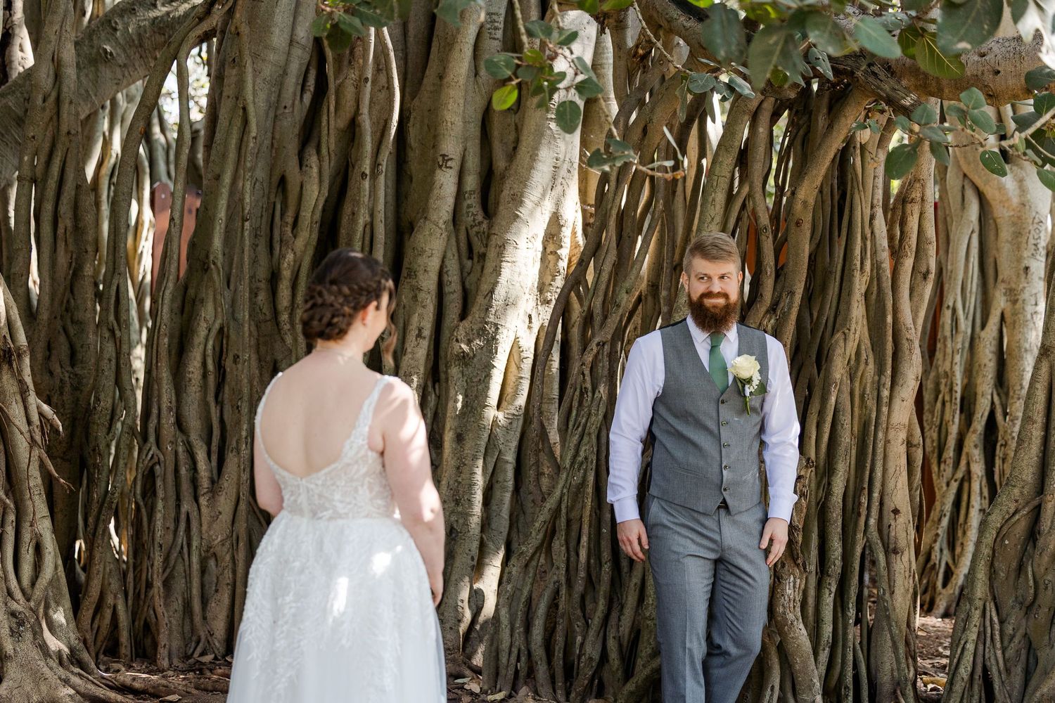 Bride and groom share a first look moment beneath a sprawling banyan tree, bride in lace gown, groom in gray vest.
