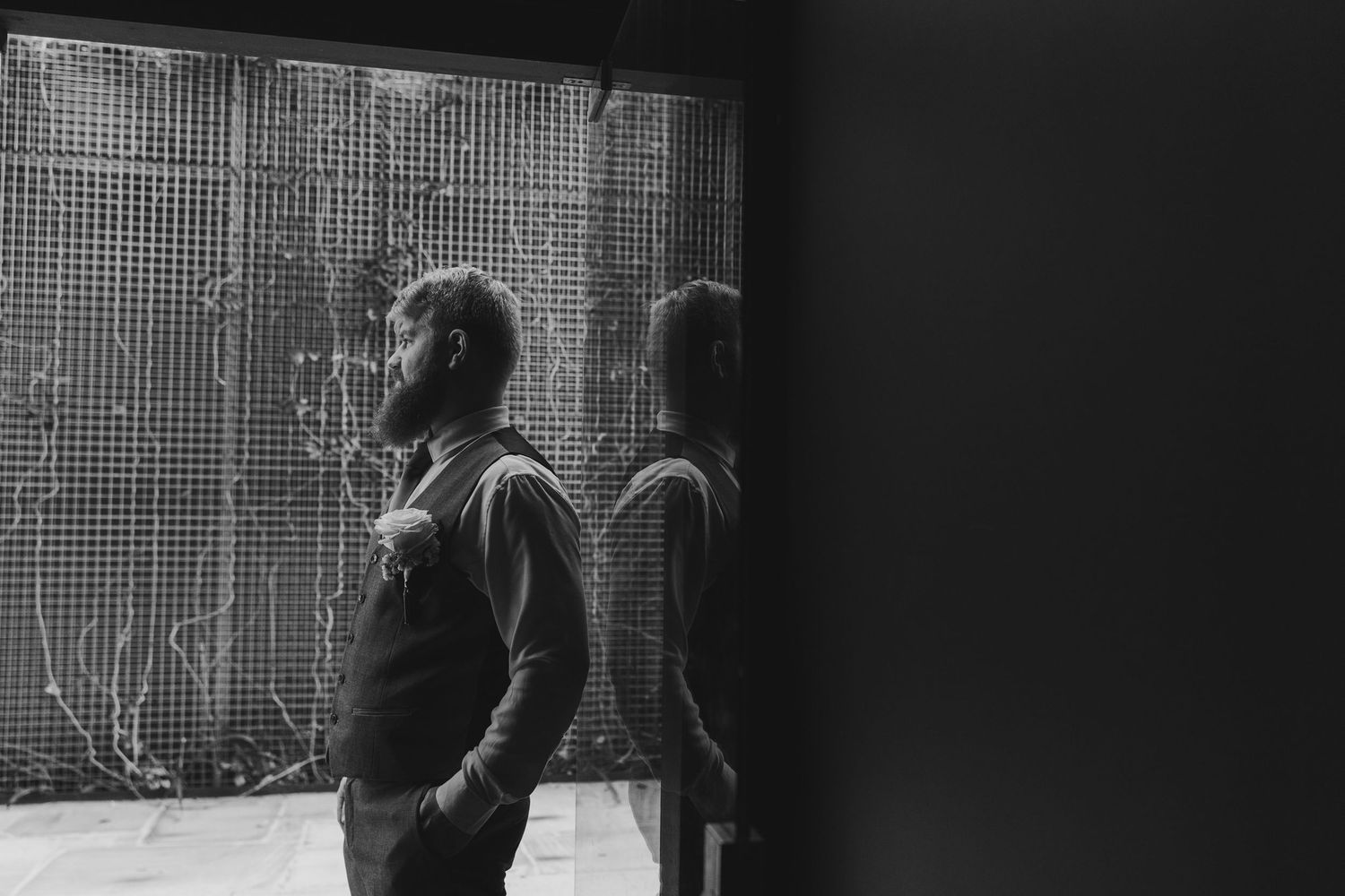 Black and white photo of a bearded man standing thoughtfully before a large textured window, with his reflection visible.