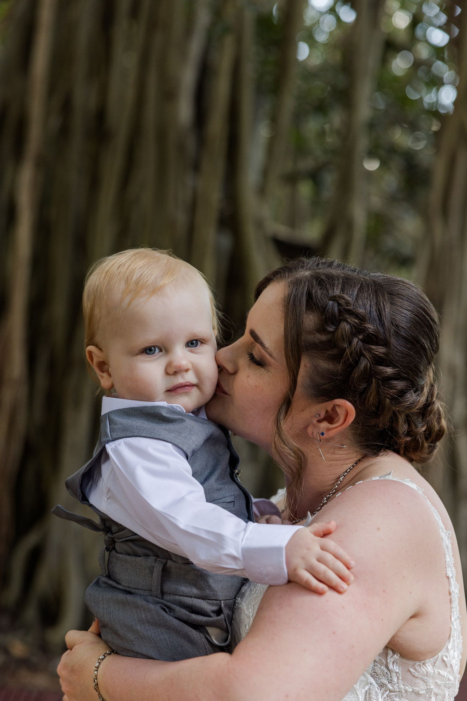 Bride in white lace gown kissing a baby boy in gray vest suit, outdoors near large tree roots.