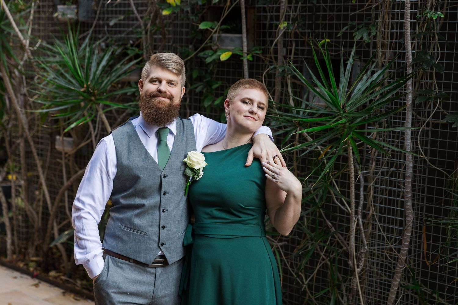 A bearded groom in a gray vest and green tie poses with a woman in an emerald green dress at an outdoor wedding.
